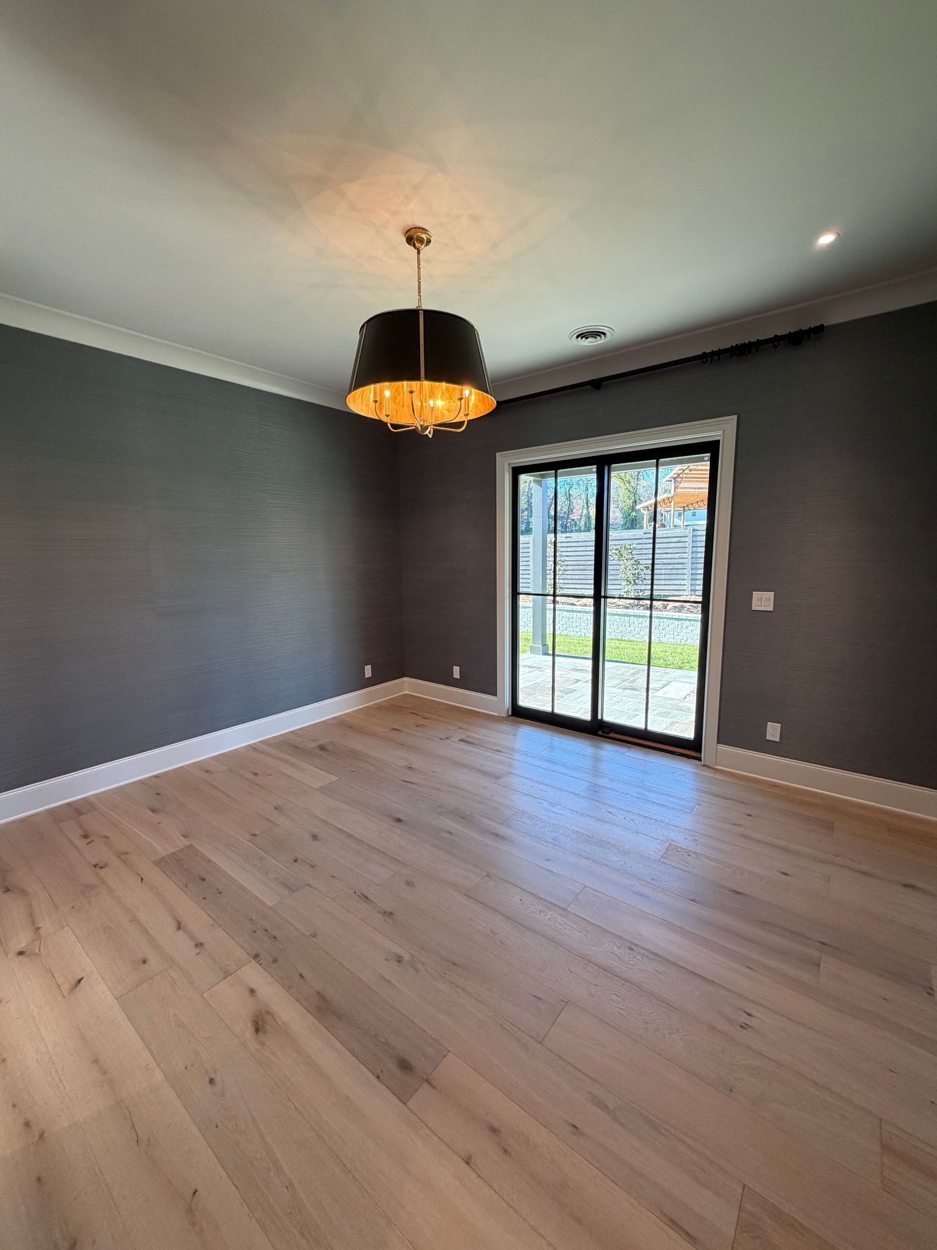 Empty modern room with gray walls, light wood floors, and a black pendant light by sliding glass doors.