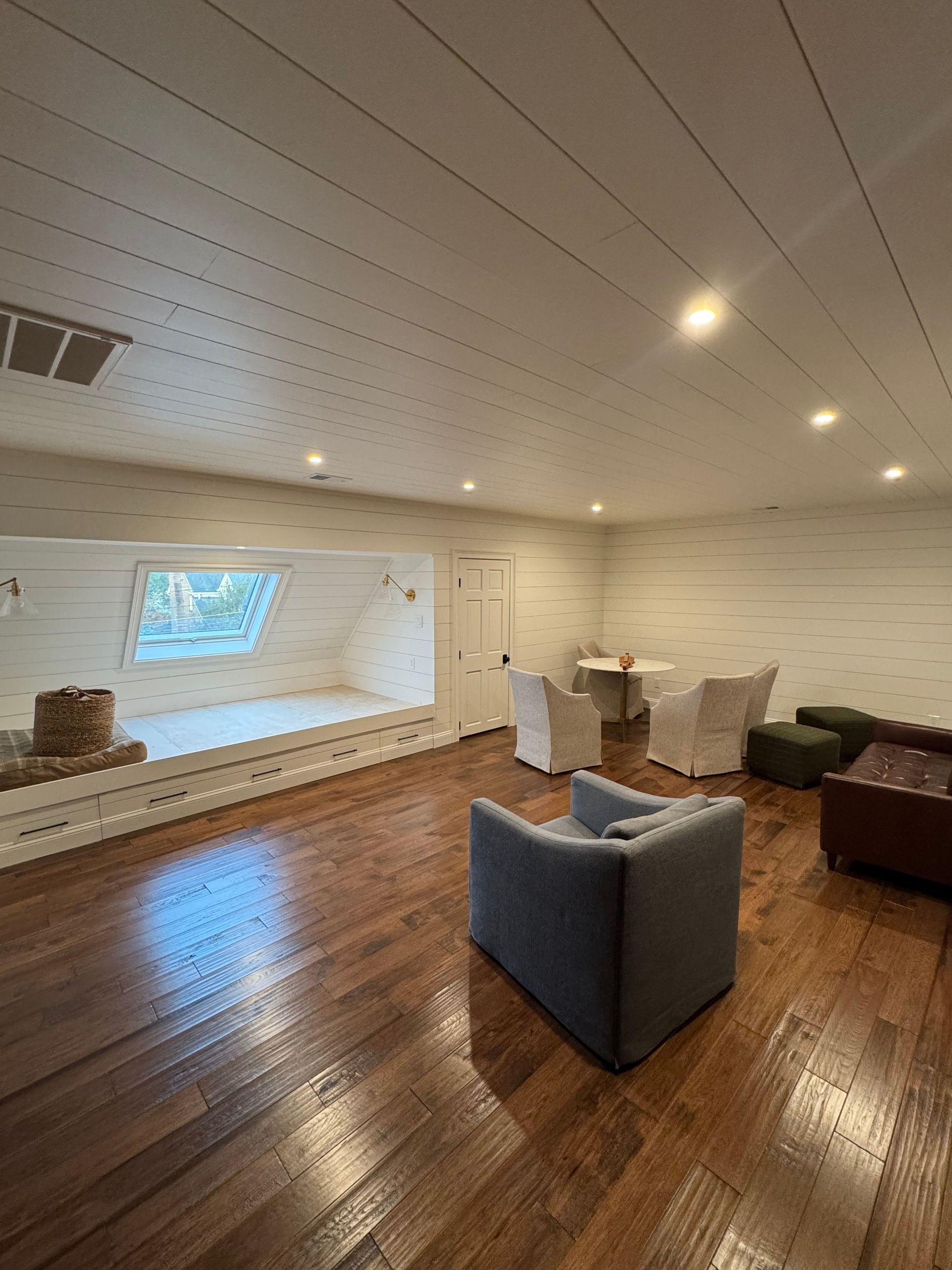 Modern living room with wood floors, white ceiling, gray chair, dining table, and wall-mounted TV at the far end