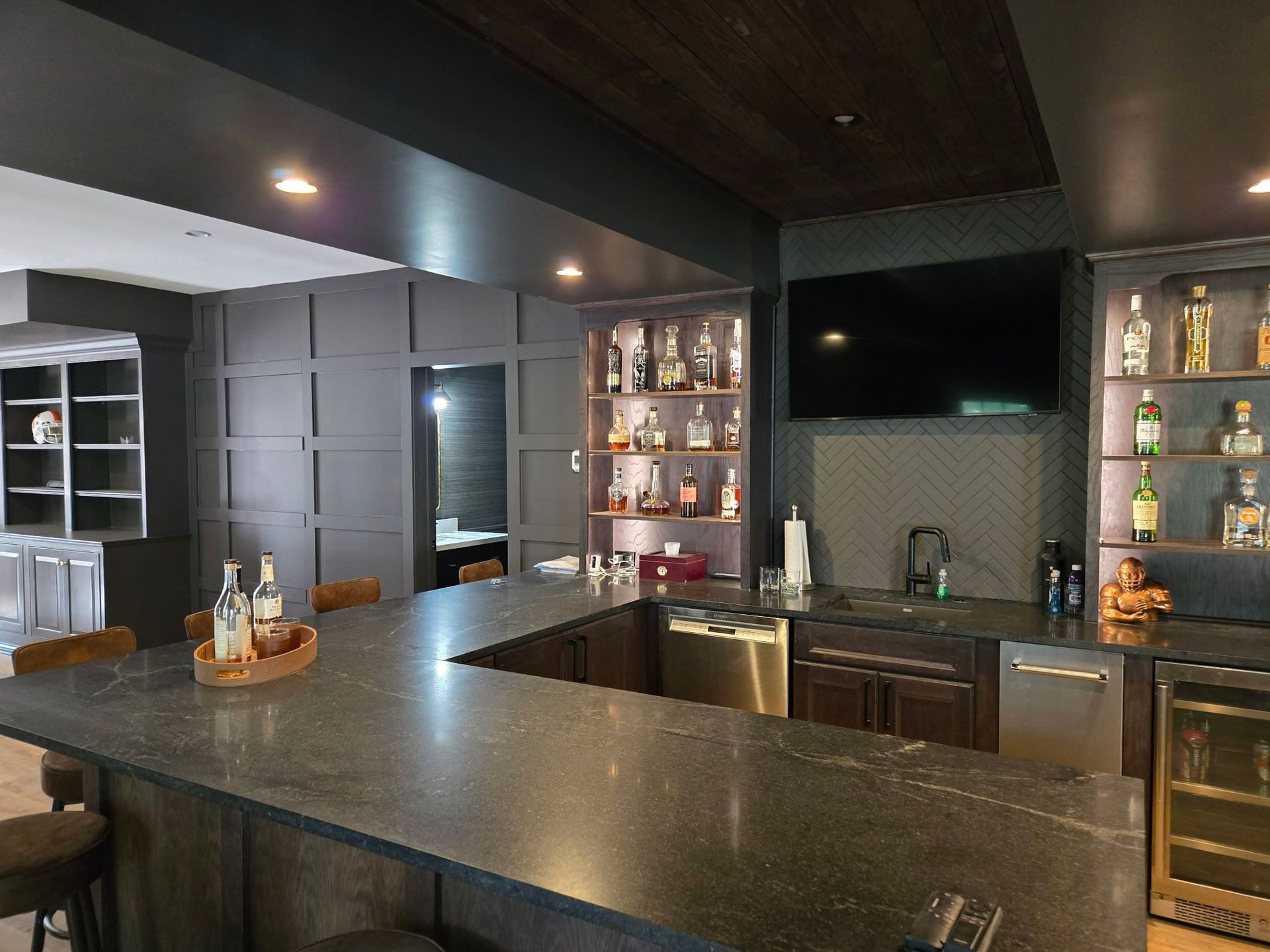 Modern home bar with granite counter, shelves of bottles, and a built-in sink under recessed lighting