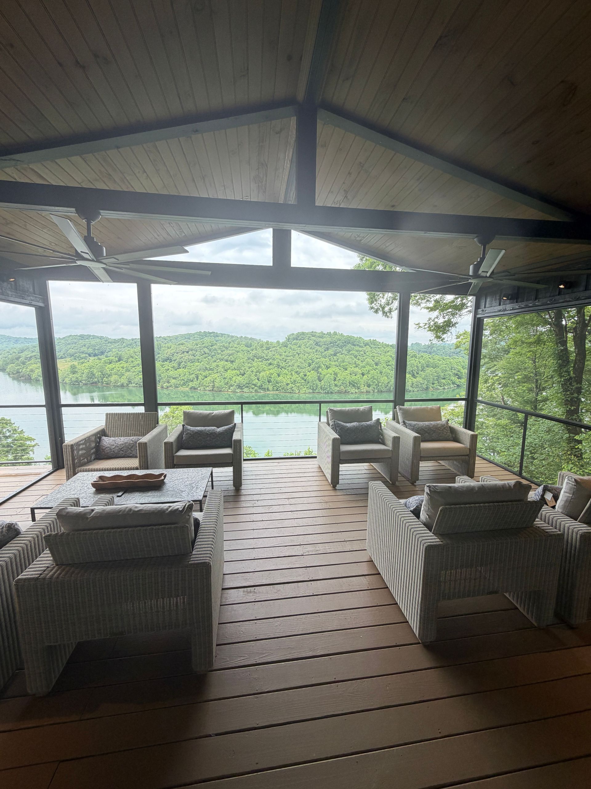 Open-air lounge with wicker sofas and chairs overlooking a green hillside and water.