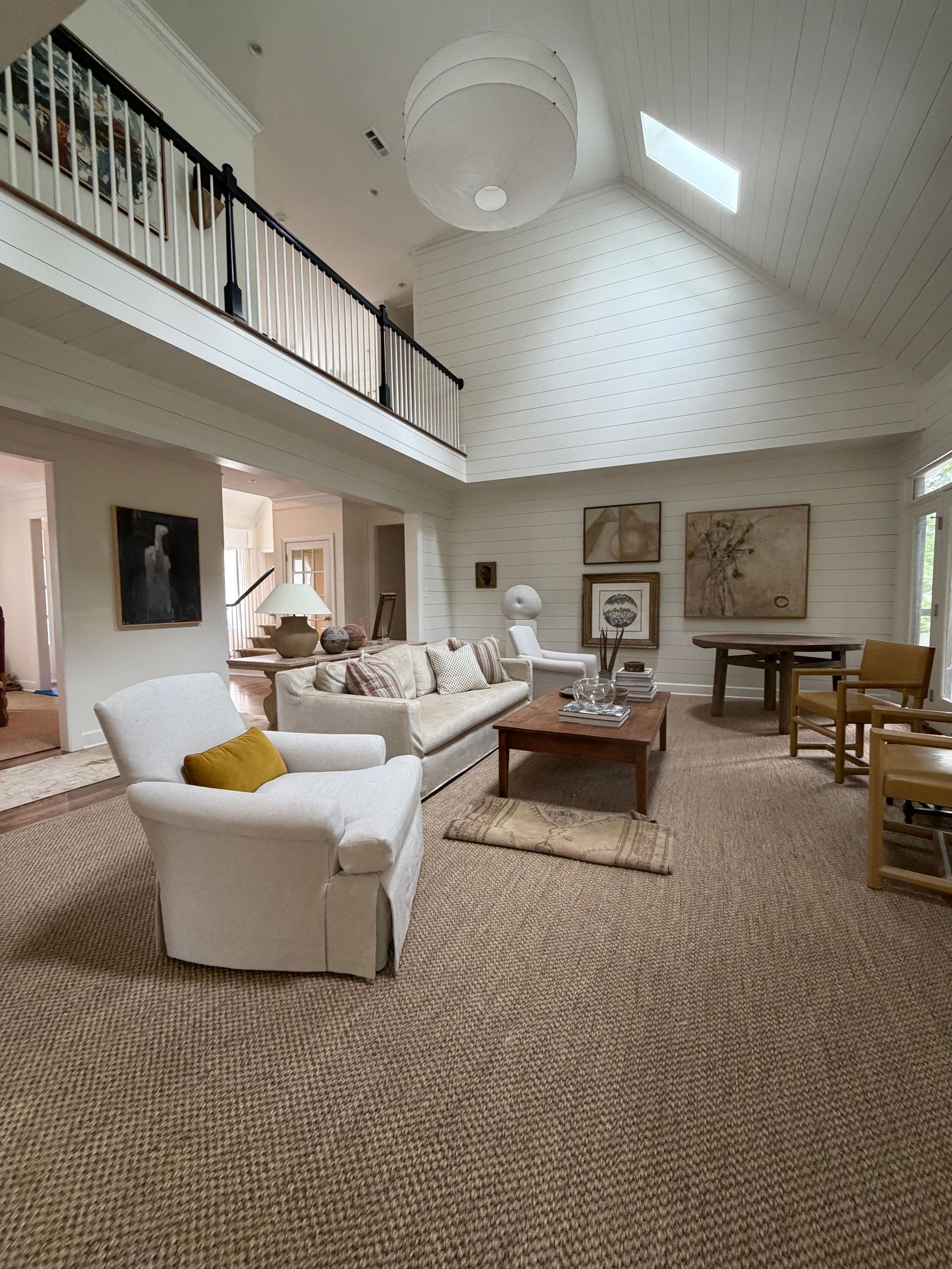 Bright living room with white sofas, woven rug, coffee table, vaulted ceiling, and balcony railing above