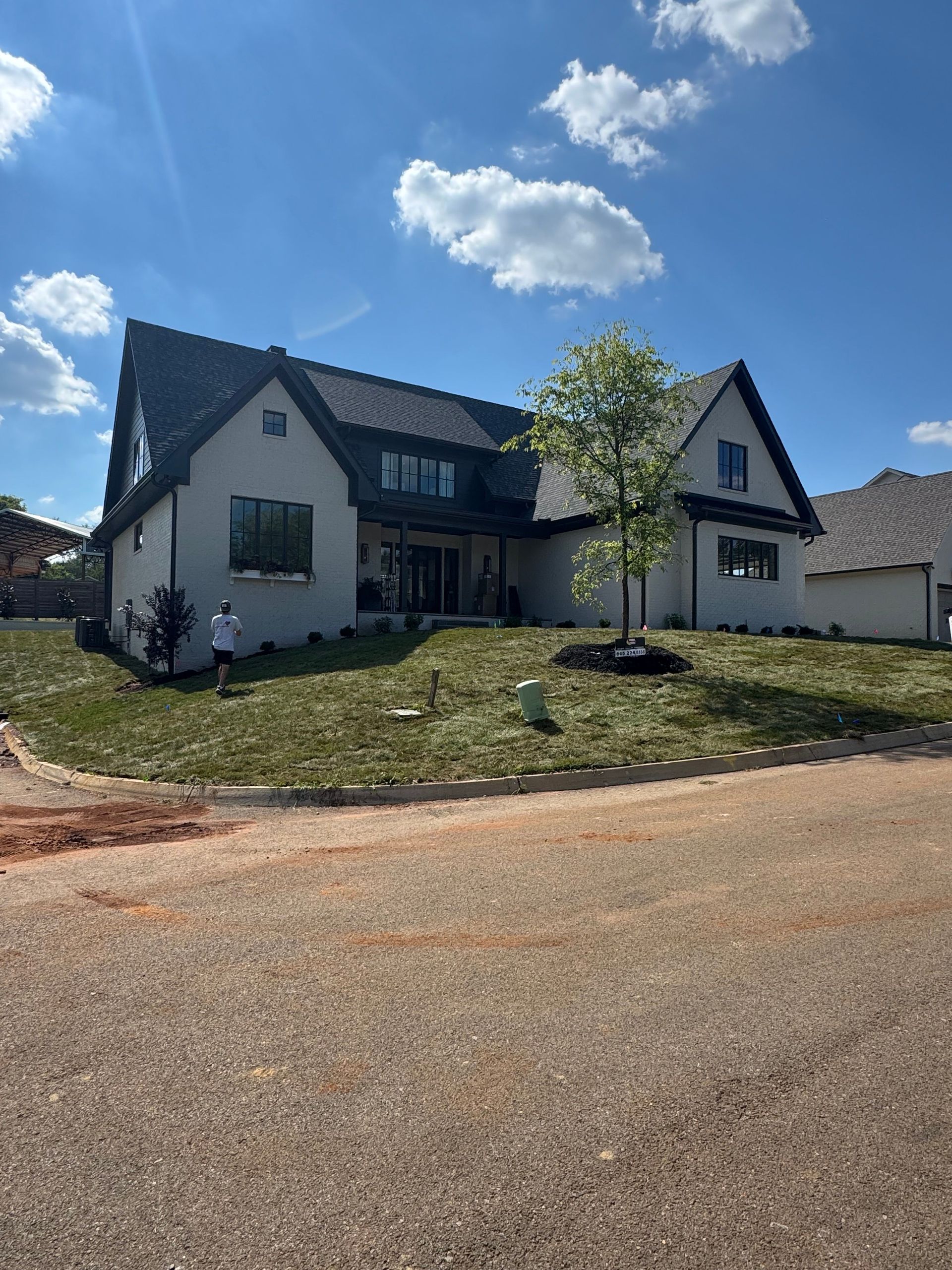 House with white siding and dark roof on a grassy hill under a blue sky, with a gravel road in front