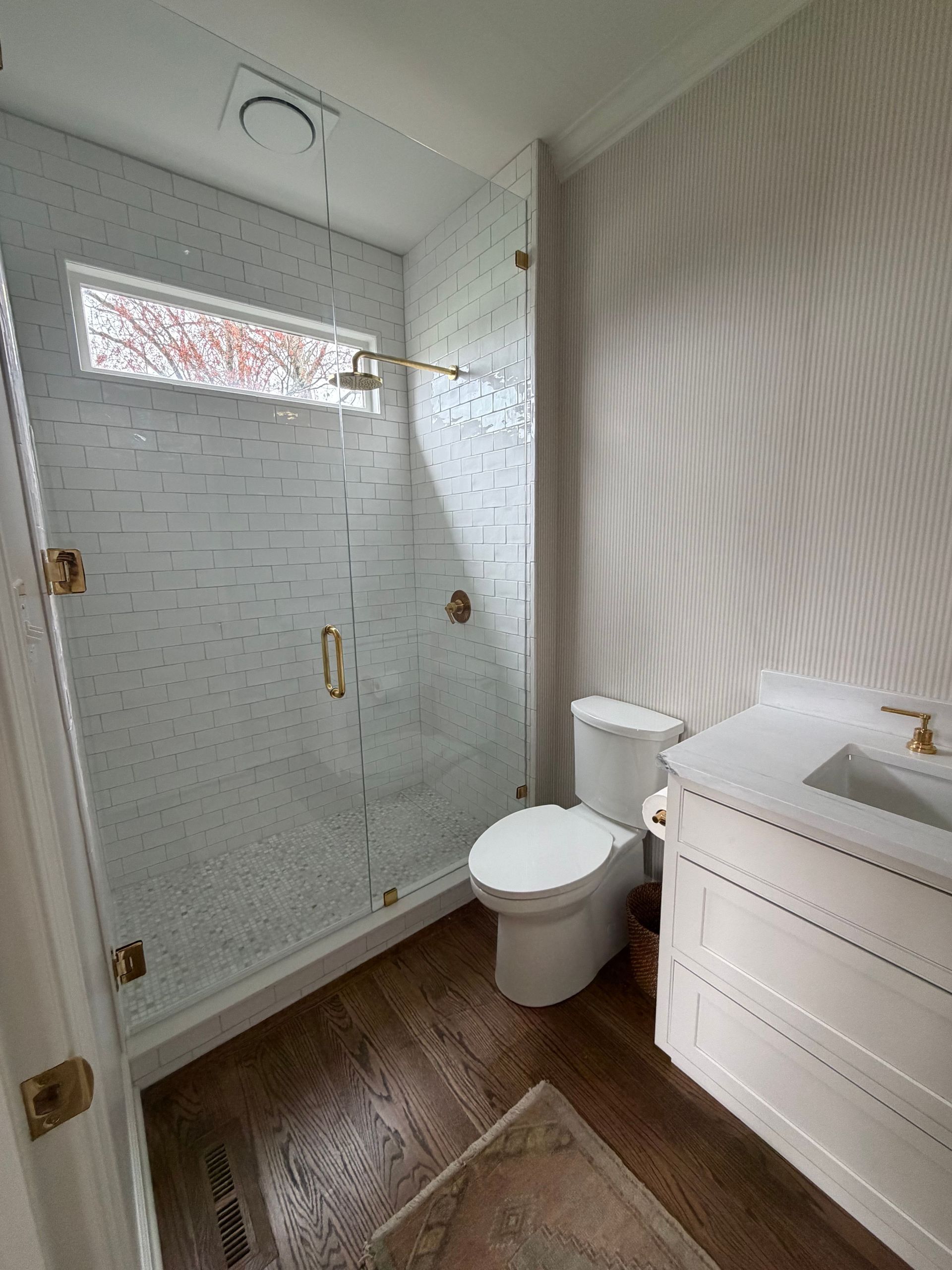 Small bathroom with glass shower, toilet, and white vanity on wood-look floor.