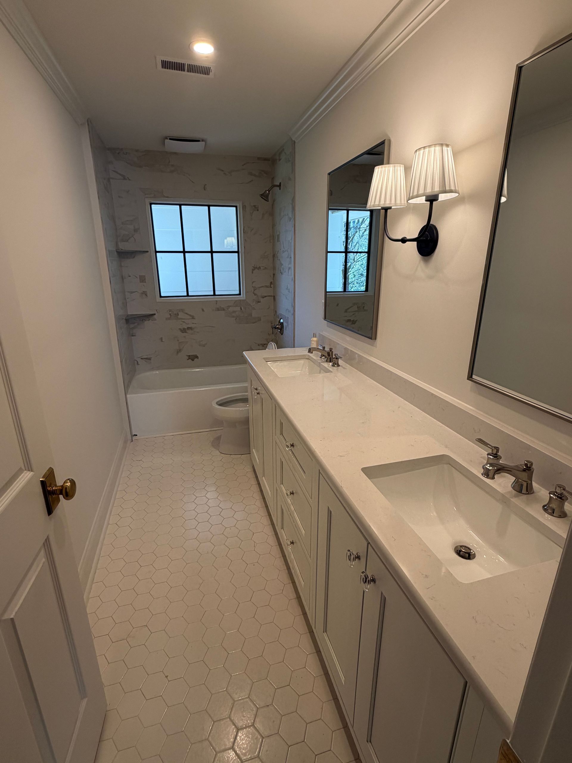 Long narrow bathroom with double vanity, large mirror, wall sconces, and a tub at the far end.