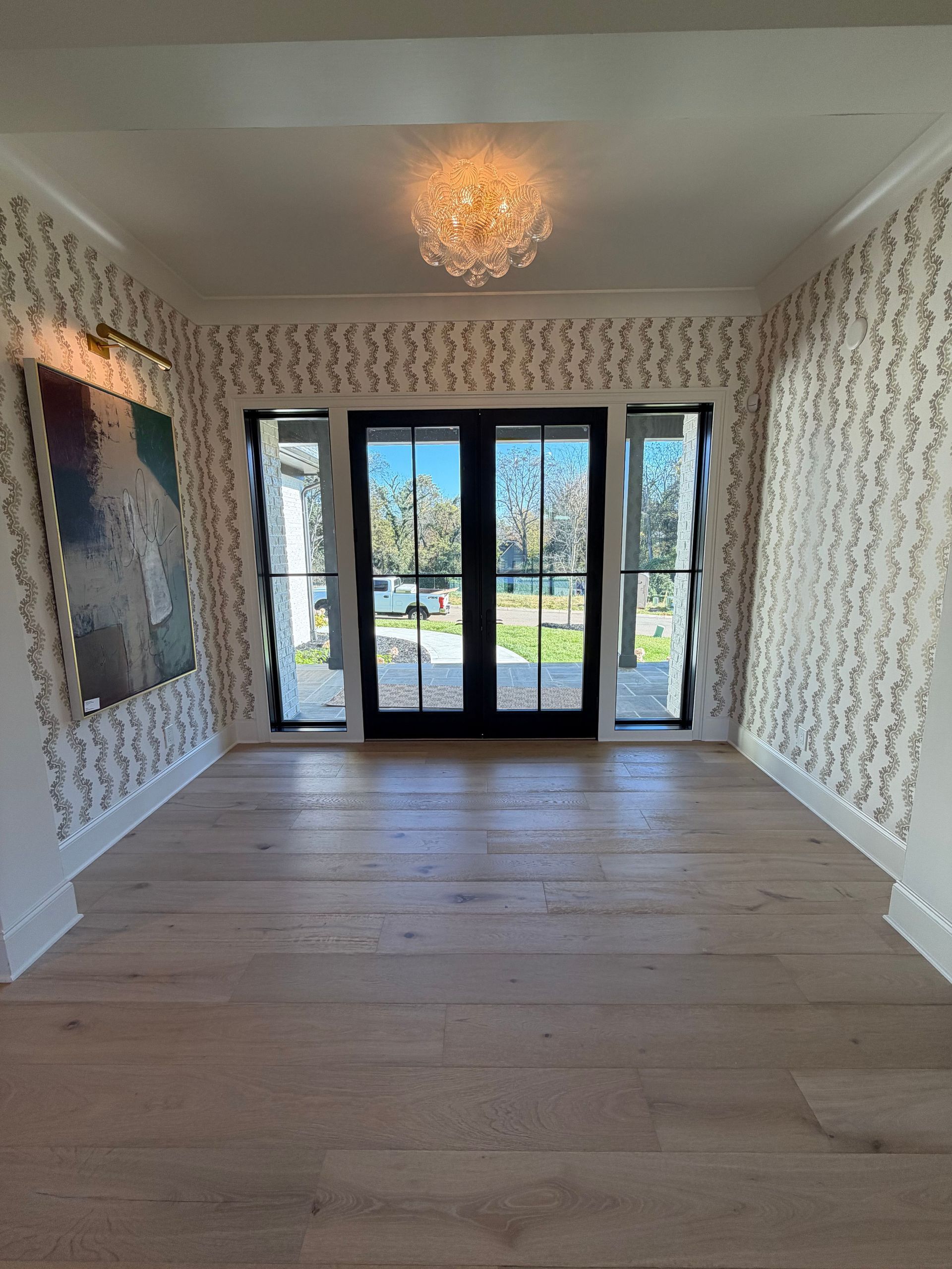 Empty elegant room with patterned wallpaper, wood floor, chandelier, and glass doors opening to a garden.