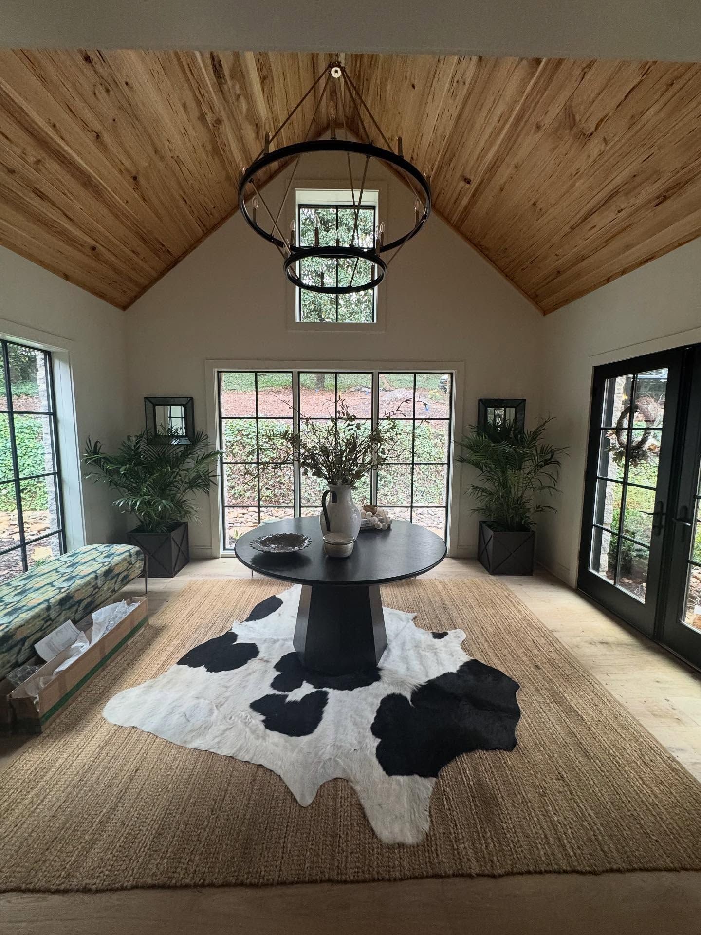 Bright sunroom with a round table on a cowhide rug, wood ceiling, and large windows overlooking greenery