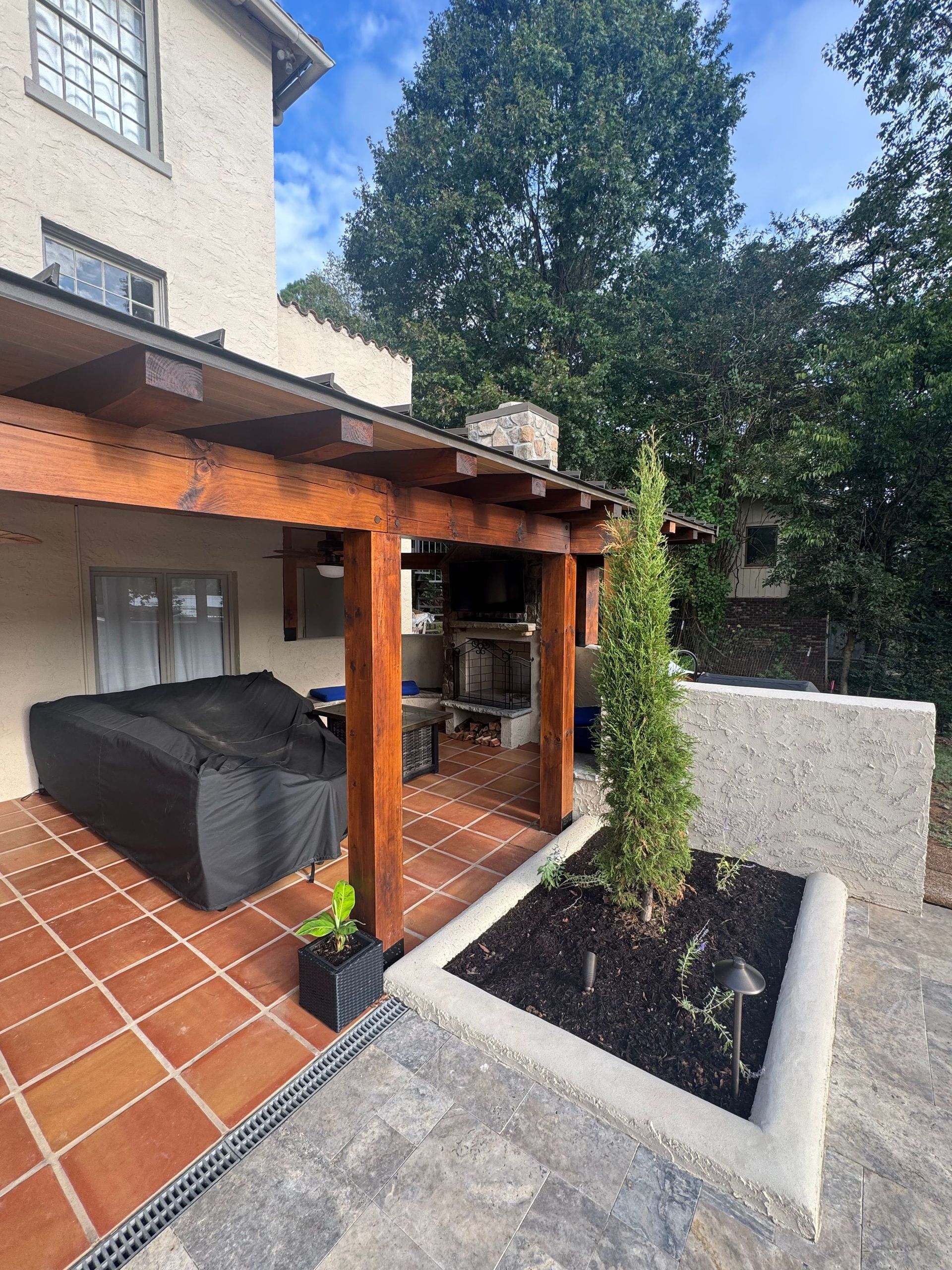 Covered patio with black sofa, tiled floor, and raised planter beside a house.