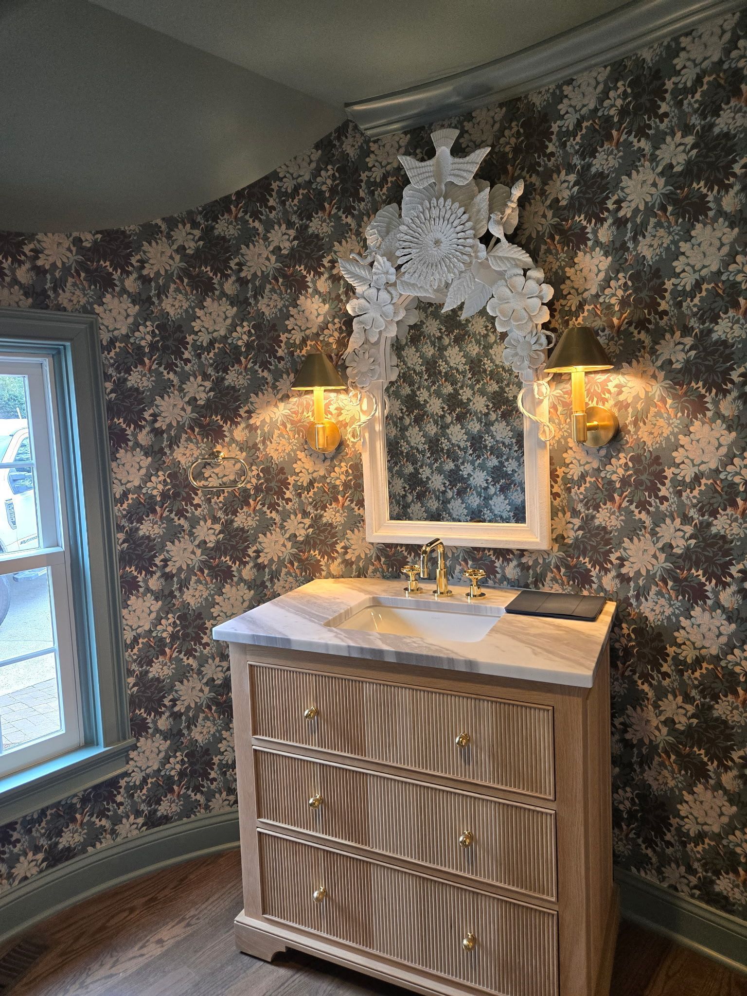 Ornate bedroom dresser with mirror and lamps against floral wallpaper, beside a window