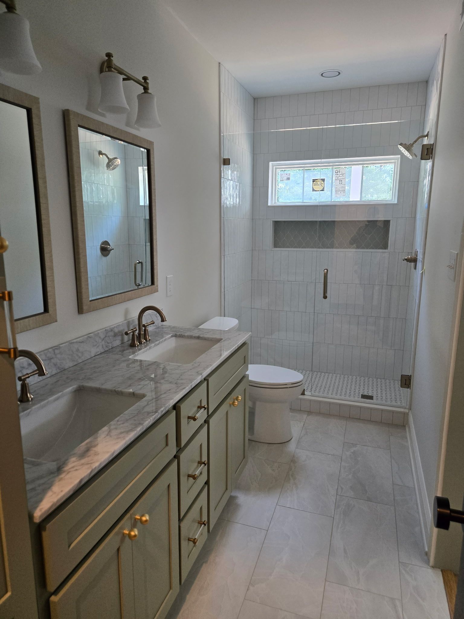 Narrow white bathroom with marble vanity, brass fixtures, mirror, toilet, and tiled shower at the far end