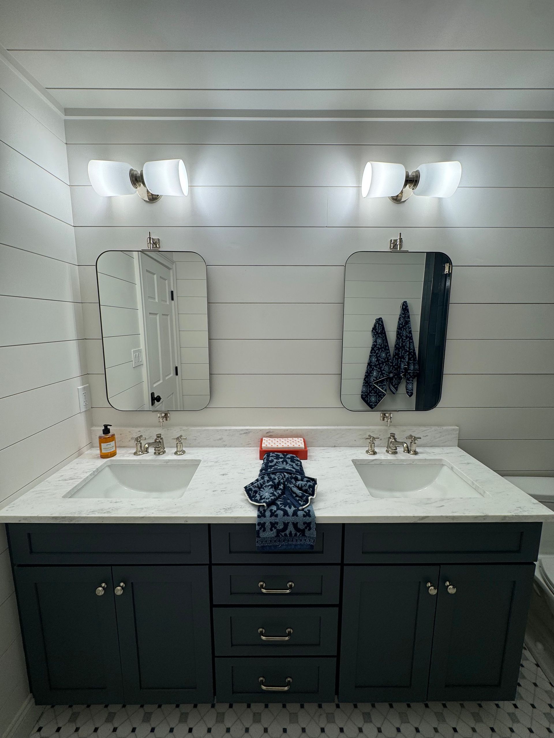 Bathroom vanity with two sinks, mirrors, wall sconces, and dark cabinets under white shiplap walls