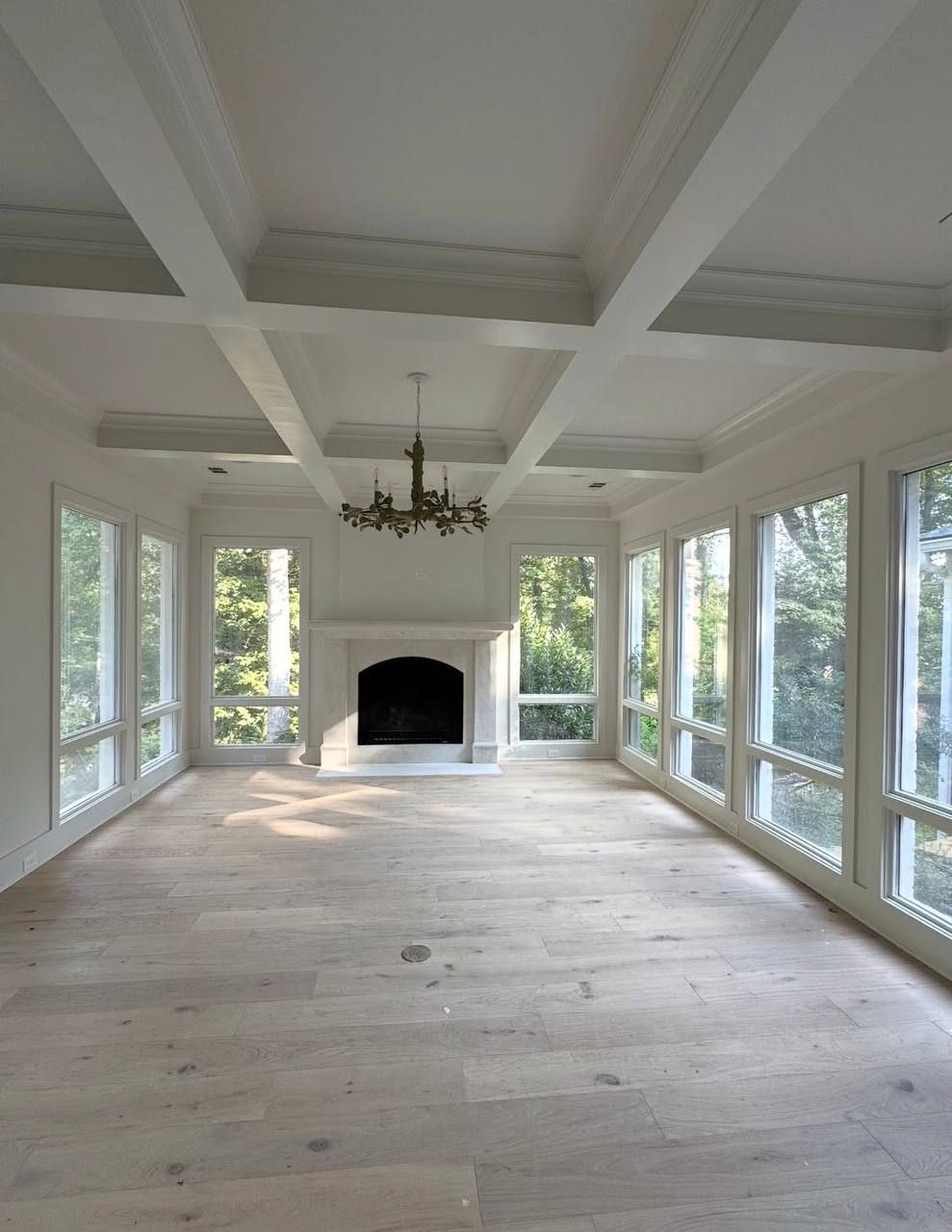 Bright sunroom with white beams, large windows, wood floor, chandelier, and fireplace.