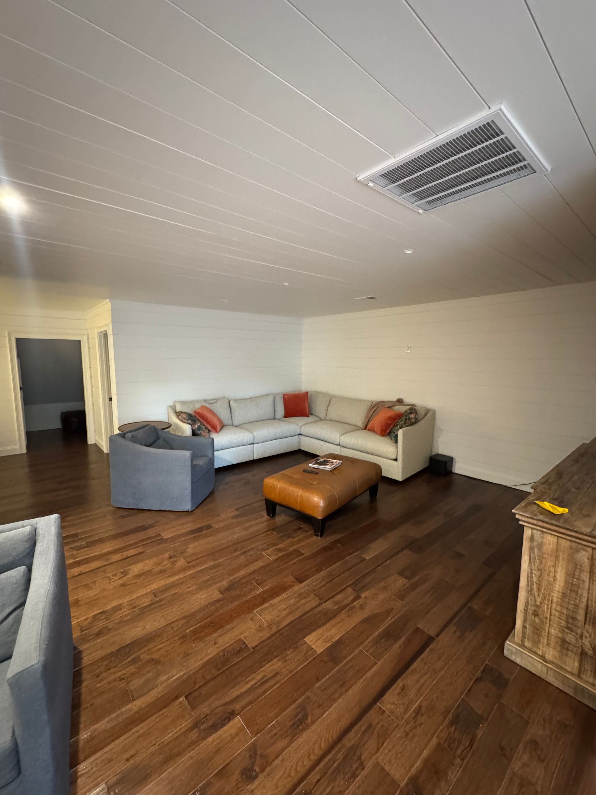 Living room with gray sectional sofa, wood floor, ottoman, and ceiling vent under a slanted white ceiling