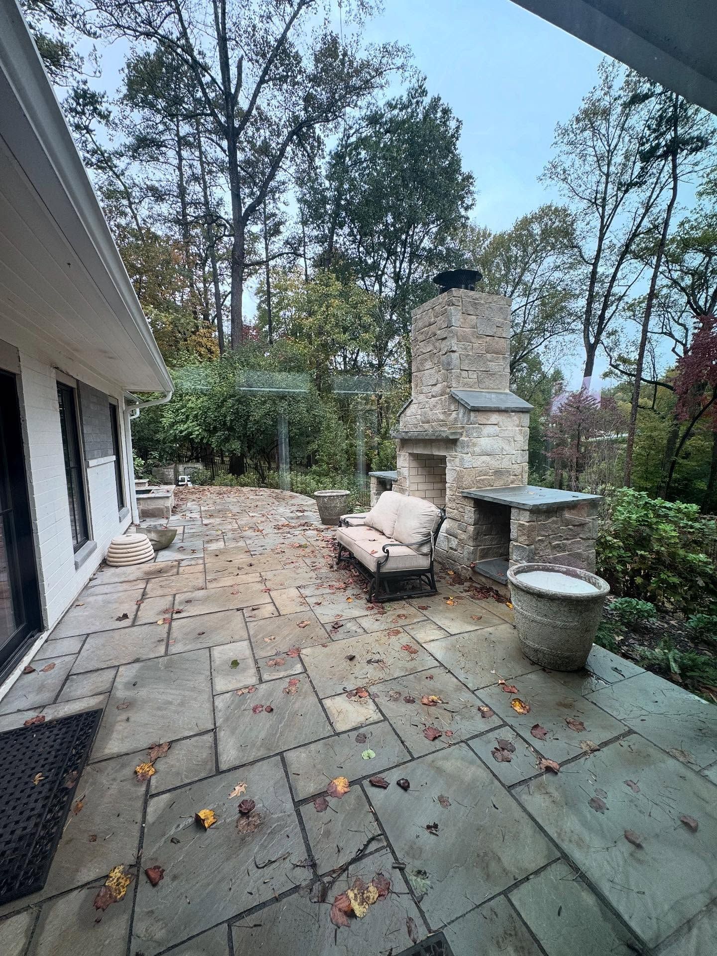 Stone patio with outdoor fireplace, lounge chair, and tall trees in a wooded backyard