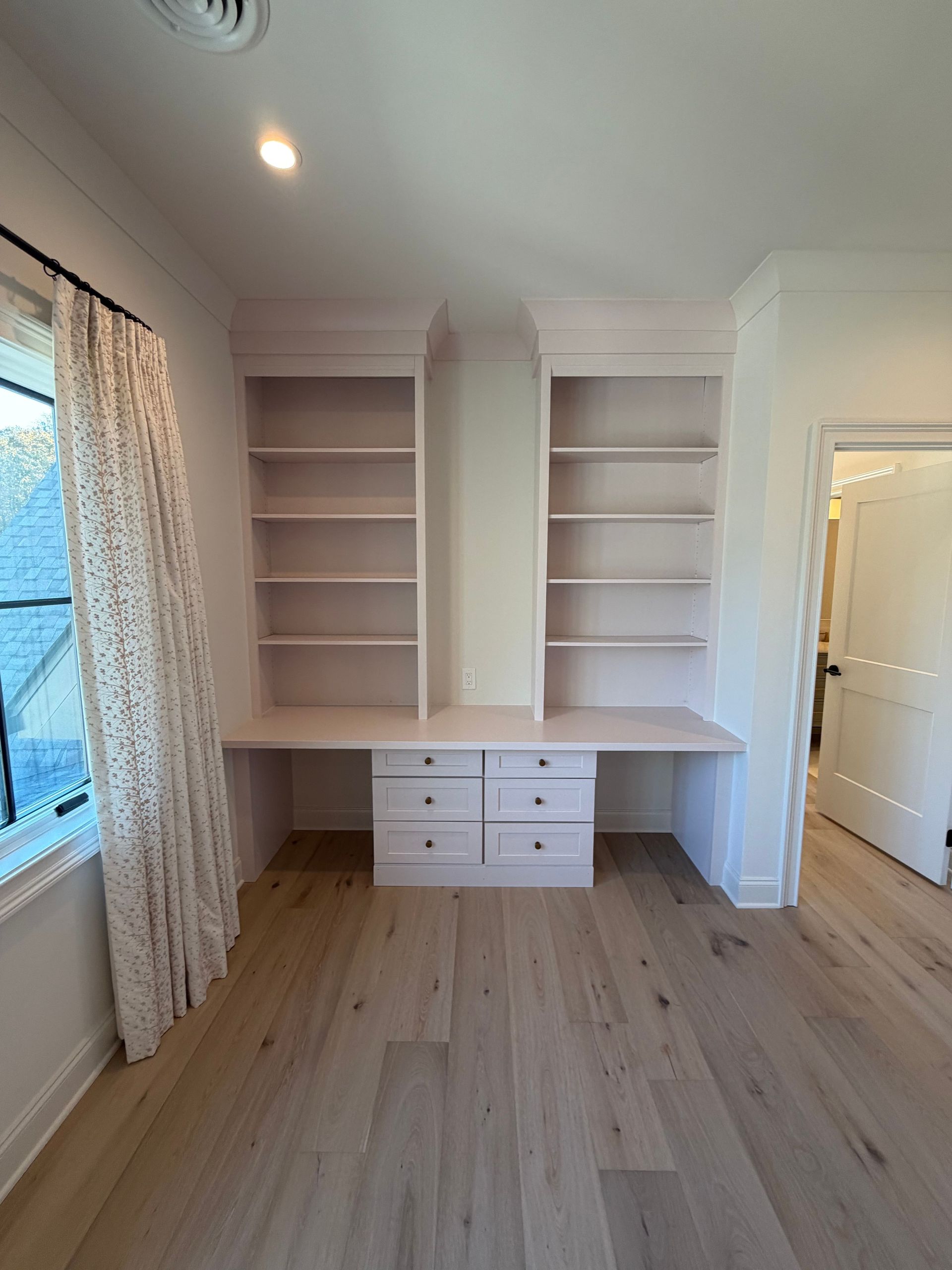 Built-in white desk with shelves and drawers in a bright, empty room with wood floors.