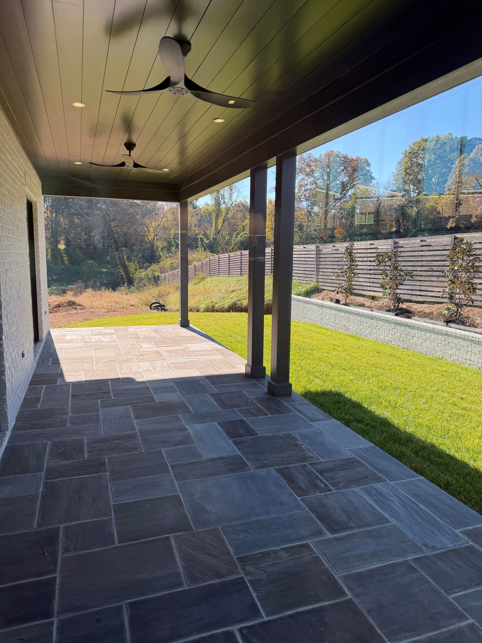Covered stone patio with ceiling fan, dark tile floor, and grassy yard bordered by trees and fencing.