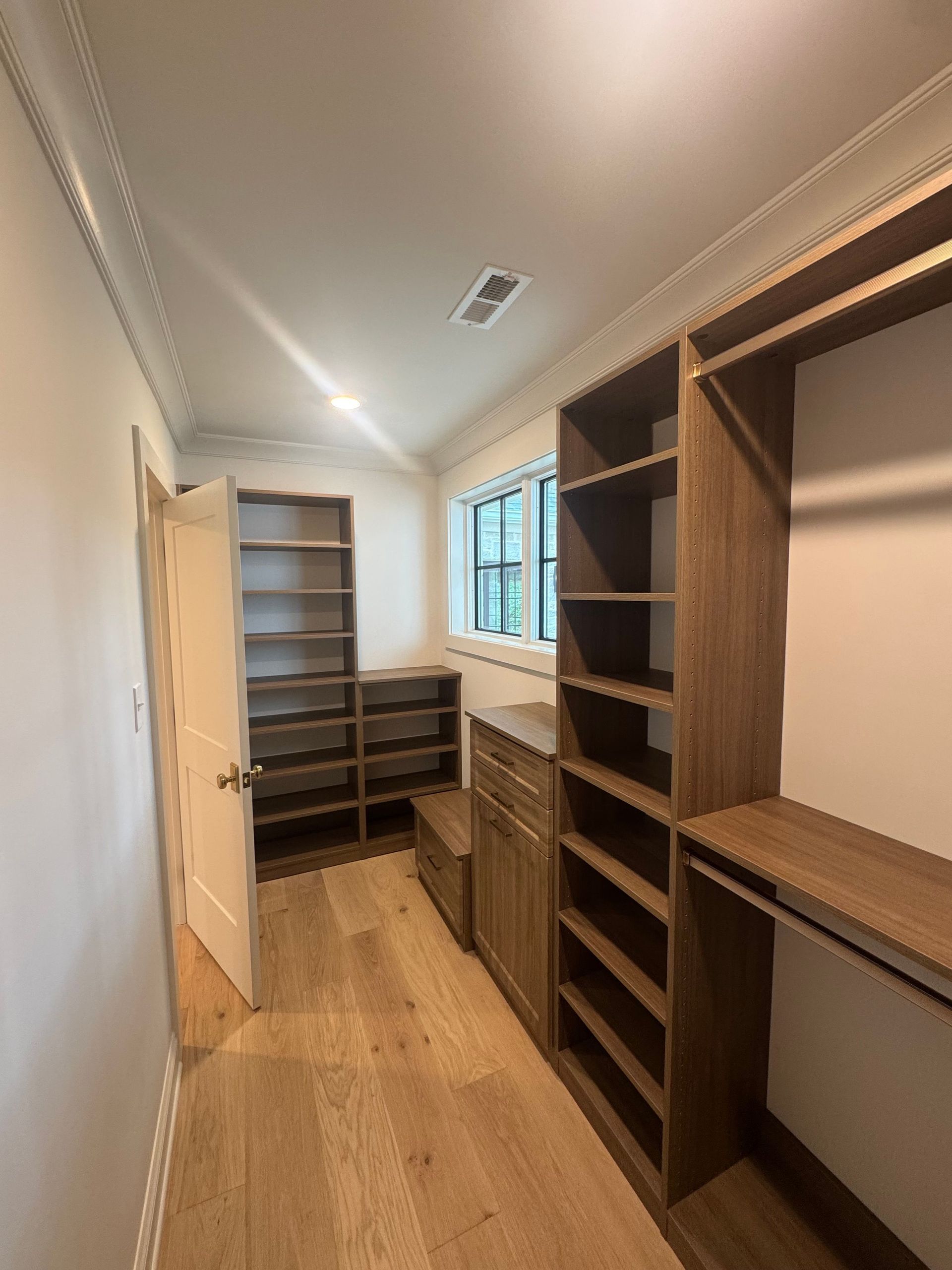 Narrow walk-in closet with built-in wooden shelves, drawers, and hanging space, lit by a window.