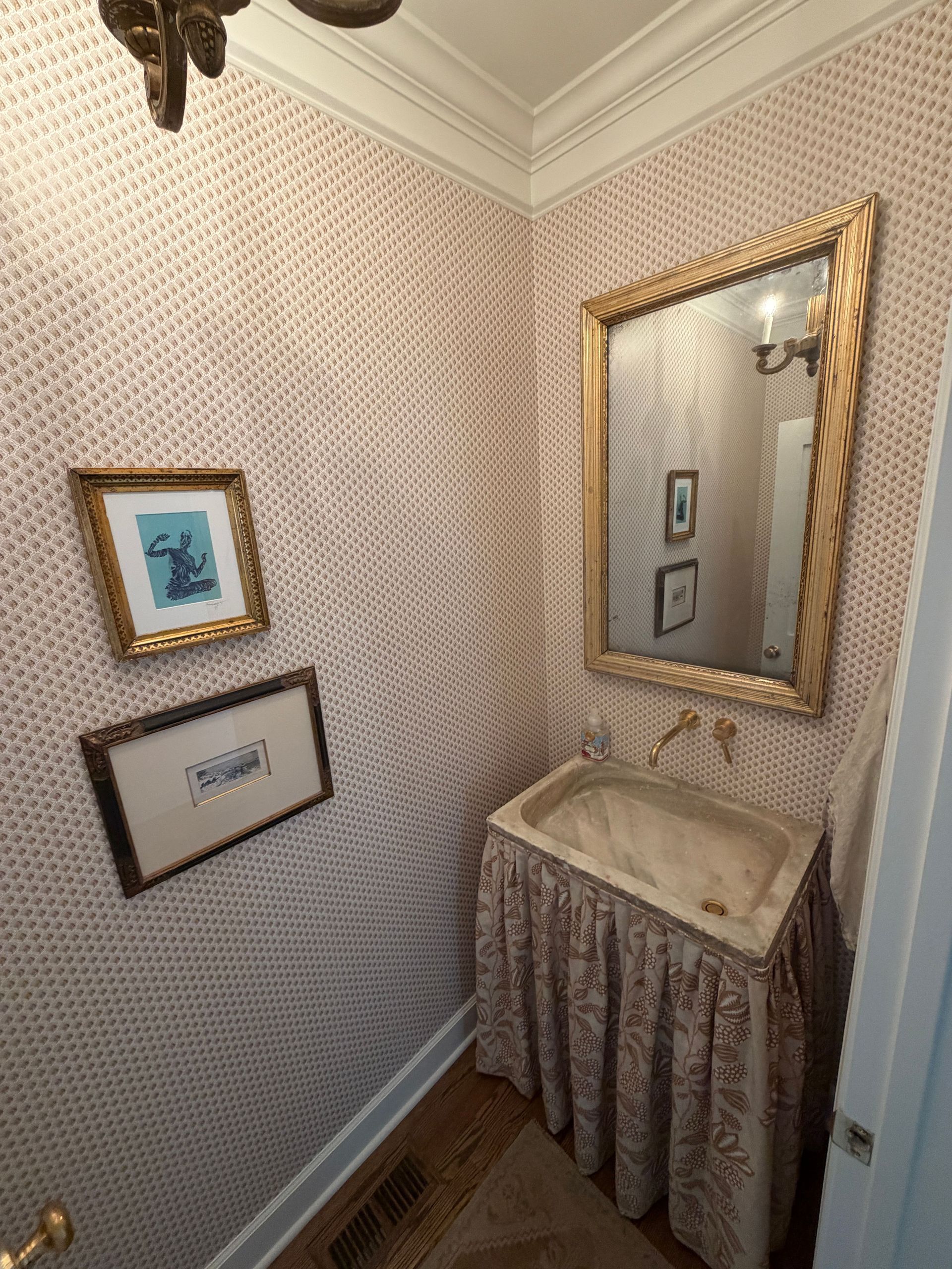 Small bathroom corner with gold mirror, patterned wallpaper, and a marble sink with a draped skirt.