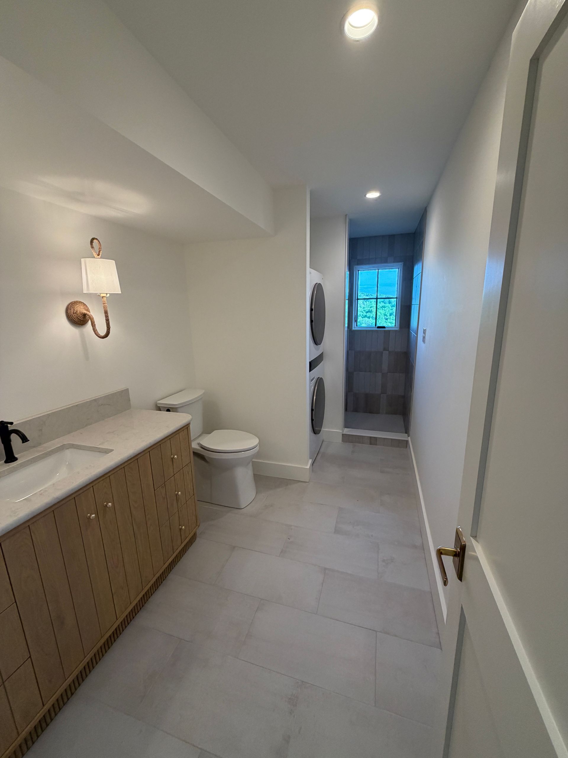 Narrow bathroom with white walls, tan vanity, toilet, and a bright window at the end of the hall.