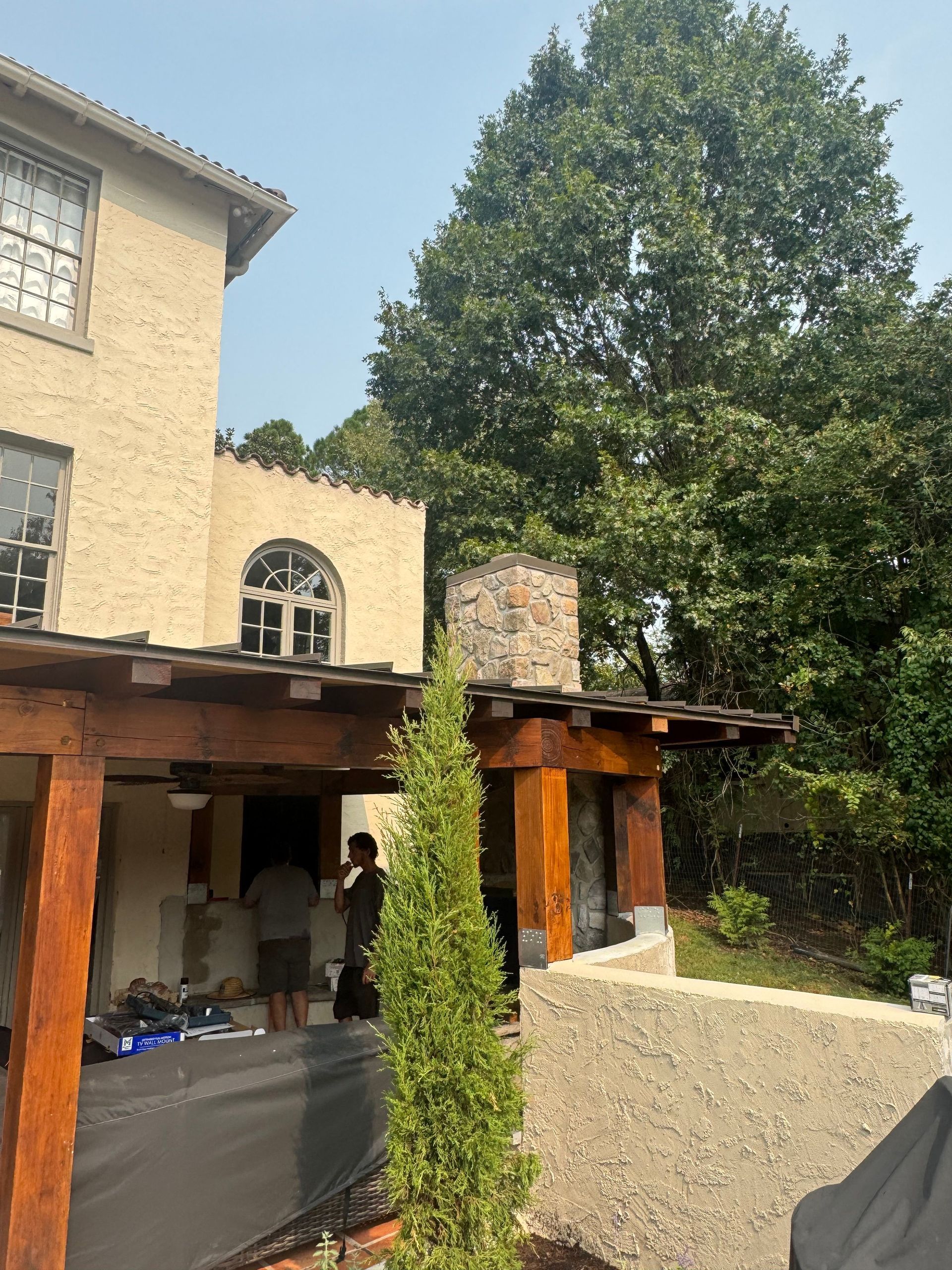Sunlit stone house patio with wooden pergola, tall cypress, and a large leafy tree in the yard