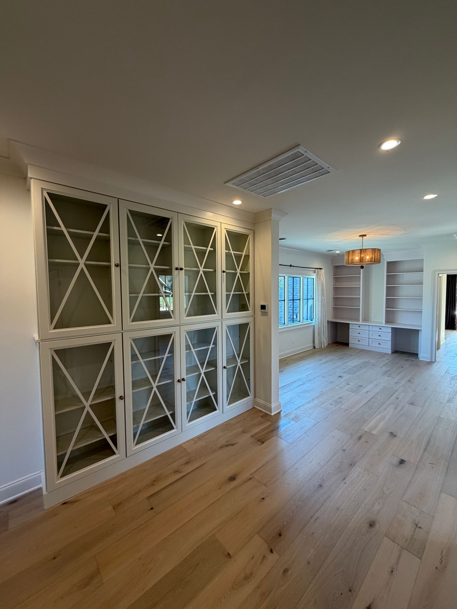 Bright open living room with hardwood floors, white built-in cabinets, and a kitchen visible in the back.