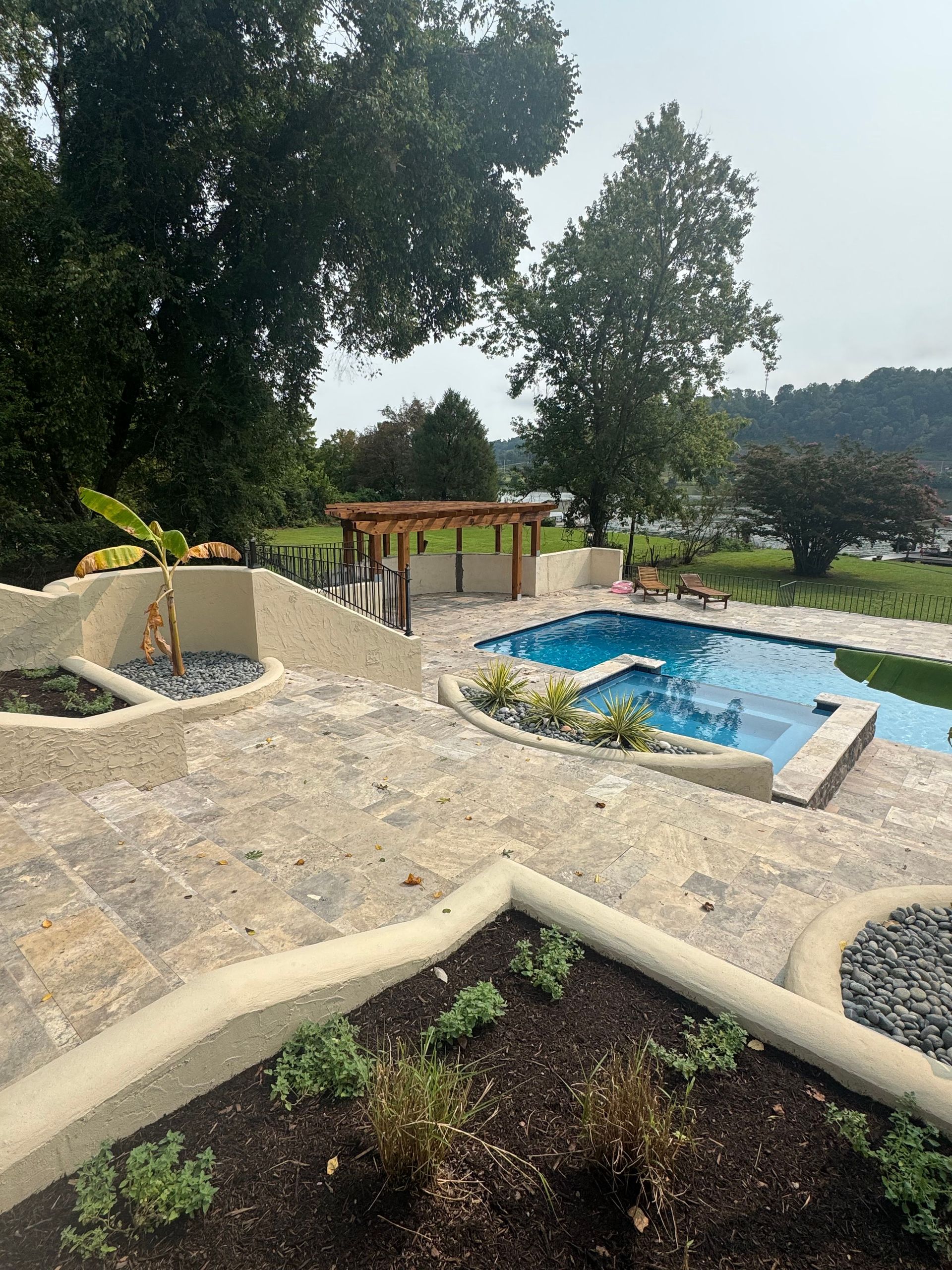 Outdoor garden with a blue pool, stone pathways, trees, and a small pergola overlooking a hillside.