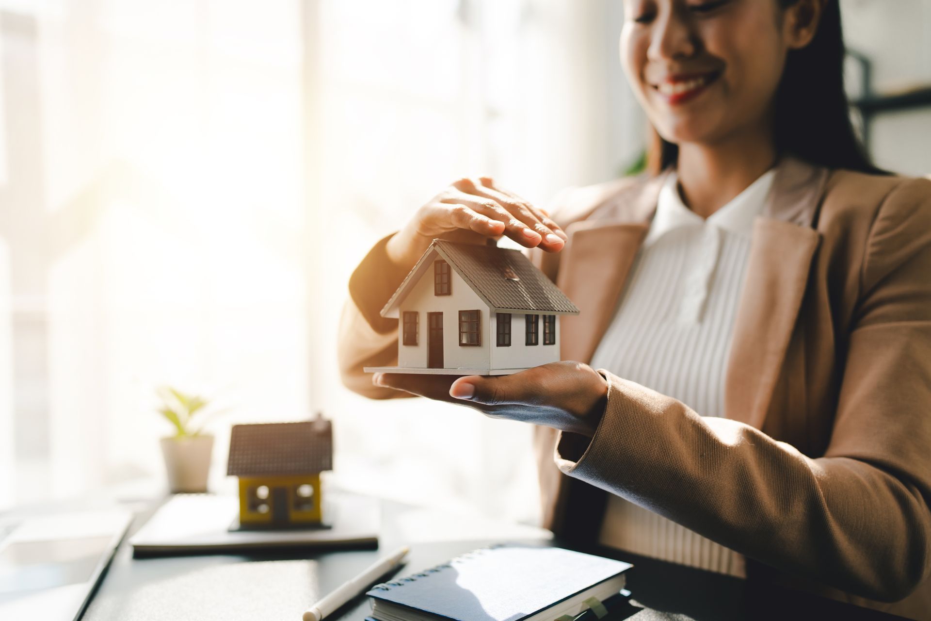 Person holding a small house model over a desk in a bright office setting
