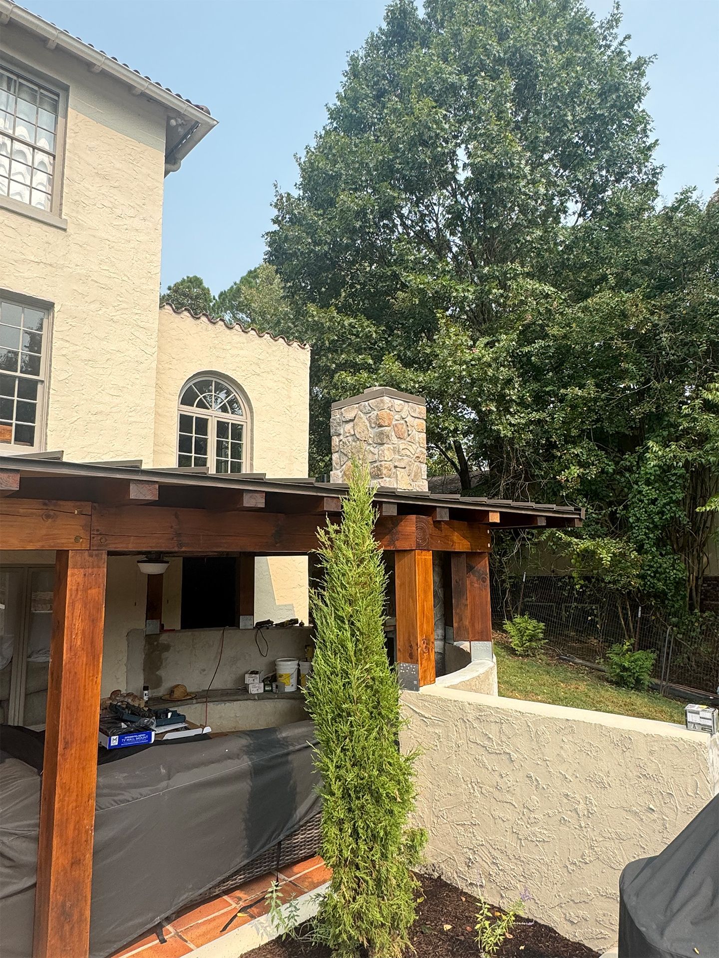 Backyard patio beside a beige house, with a tall tree, wooden pergola, and outdoor seating area