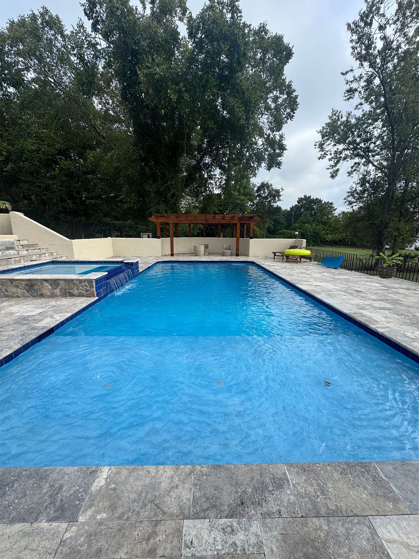 Long blue swimming pool in a stone patio with a pergola and trees beyond