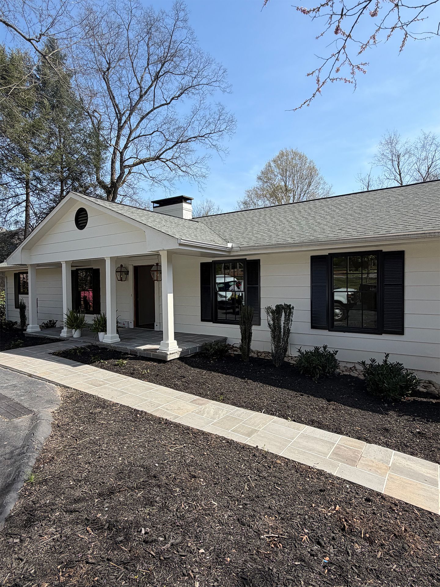 Single-story white house with dark shutters, front porch, and bare winter trees under a blue sky.