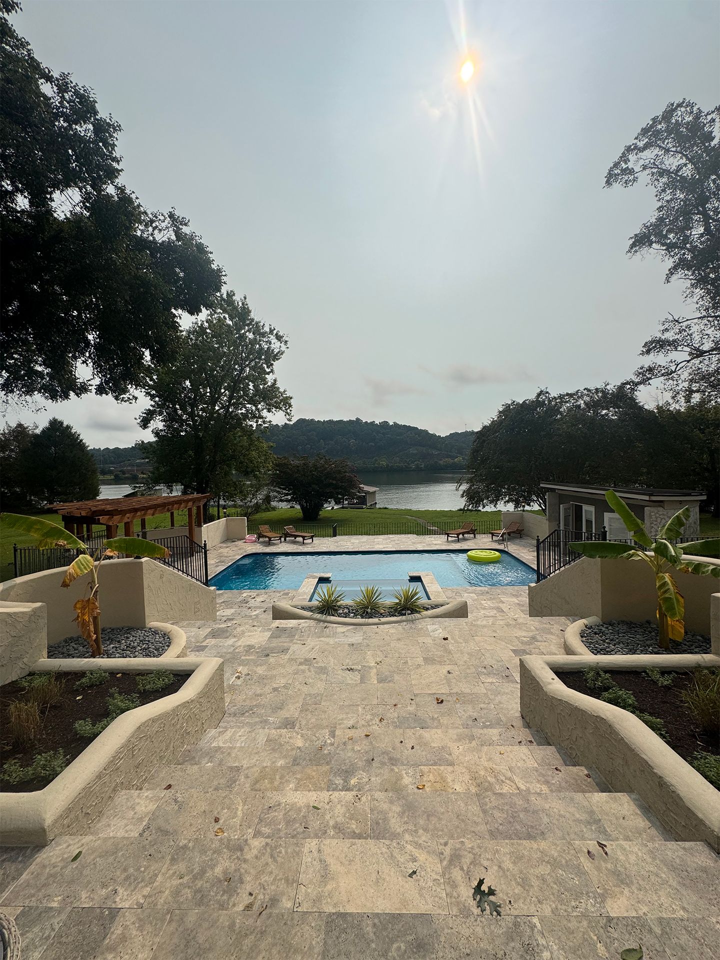 Stone steps to a pool overlooking a lake, framed by trees and planters under bright sun.