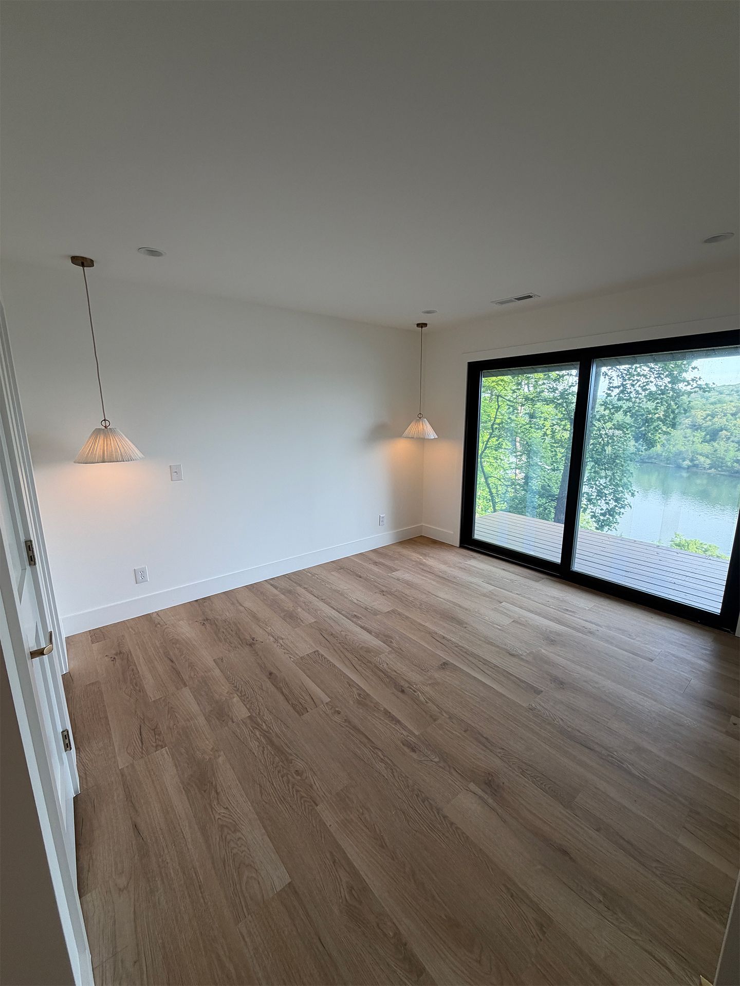 Empty modern living room with wood floors, white walls, hanging lights, and large sliding glass doors to greenery