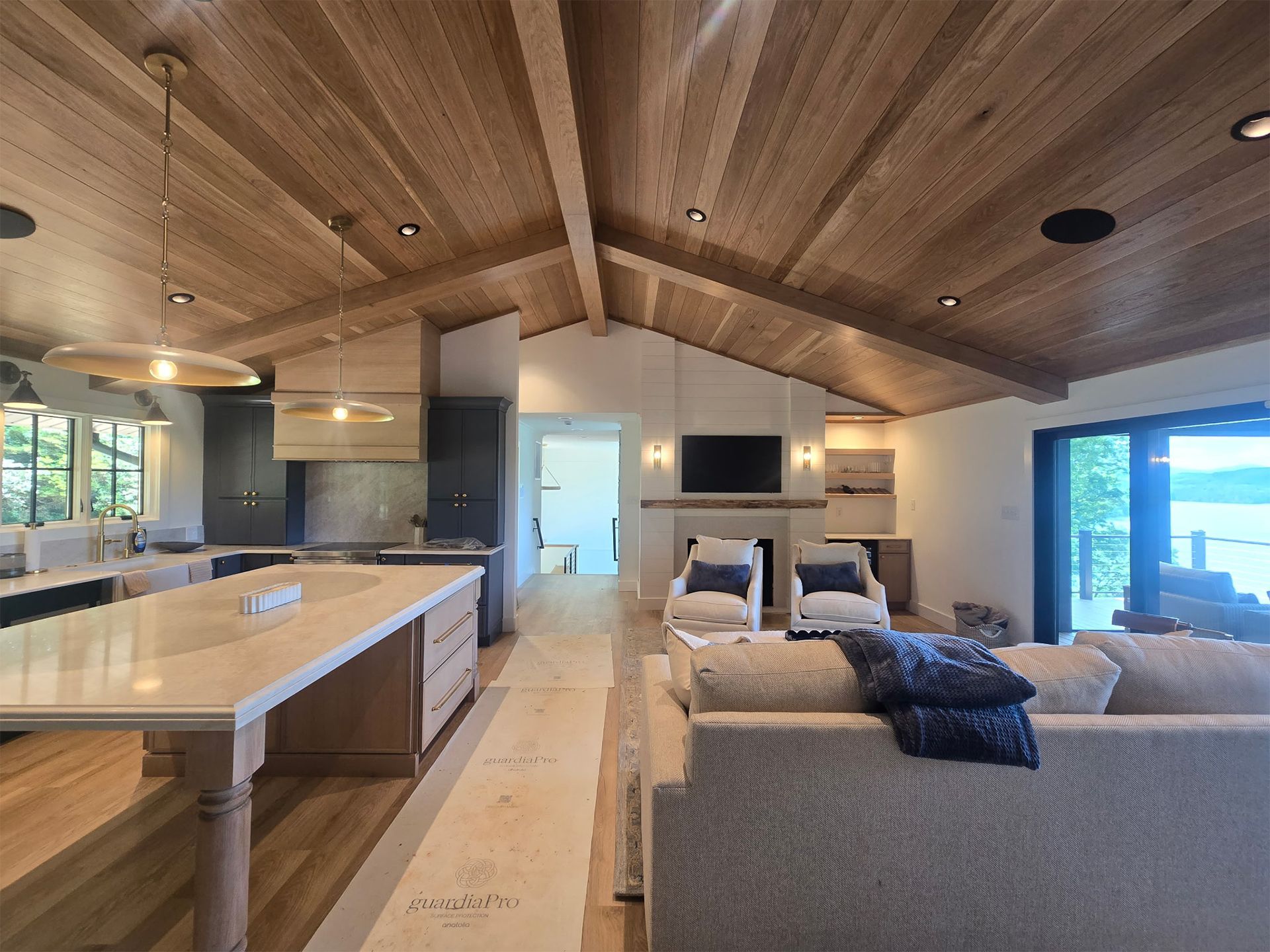 Modern open-plan living room with wood ceiling, kitchen island, and gray sectional by large windows.