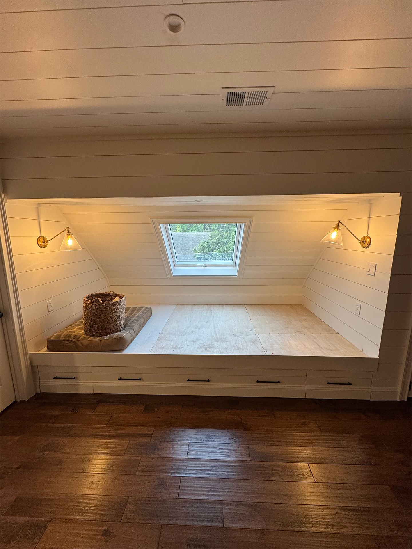 Small white-paneled alcove with a window seat, wicker basket, sconces, and a dark wooden floor