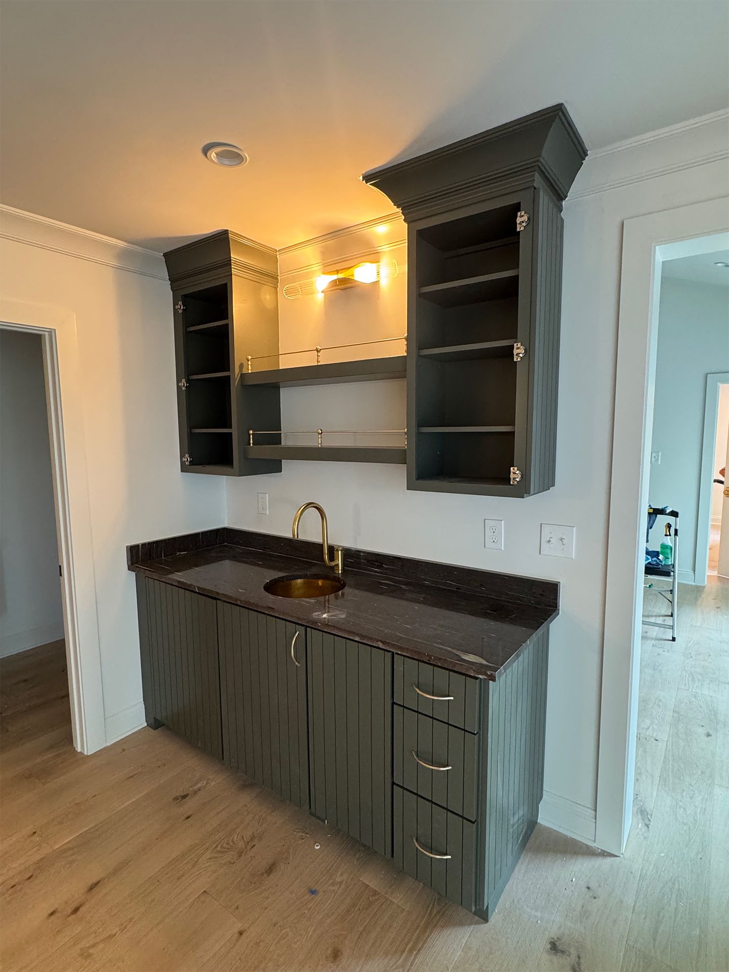Small kitchen nook with dark cabinets, black countertop, sink, and built-in wall shelves under warm lighting