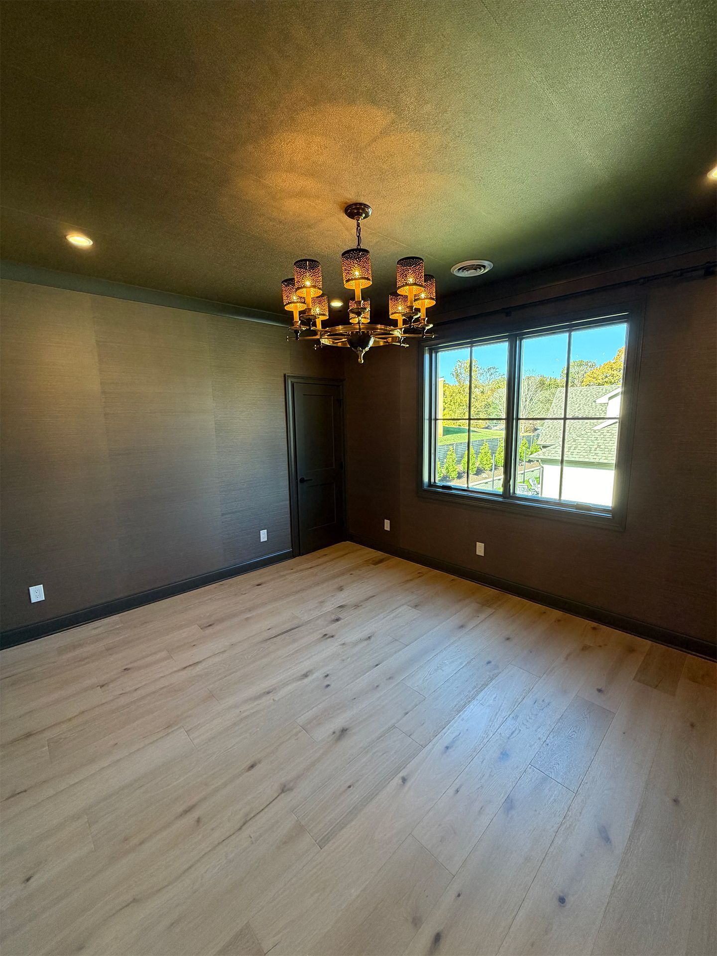 Empty room with dark walls, light wood floor, chandelier, and window showing trees outside