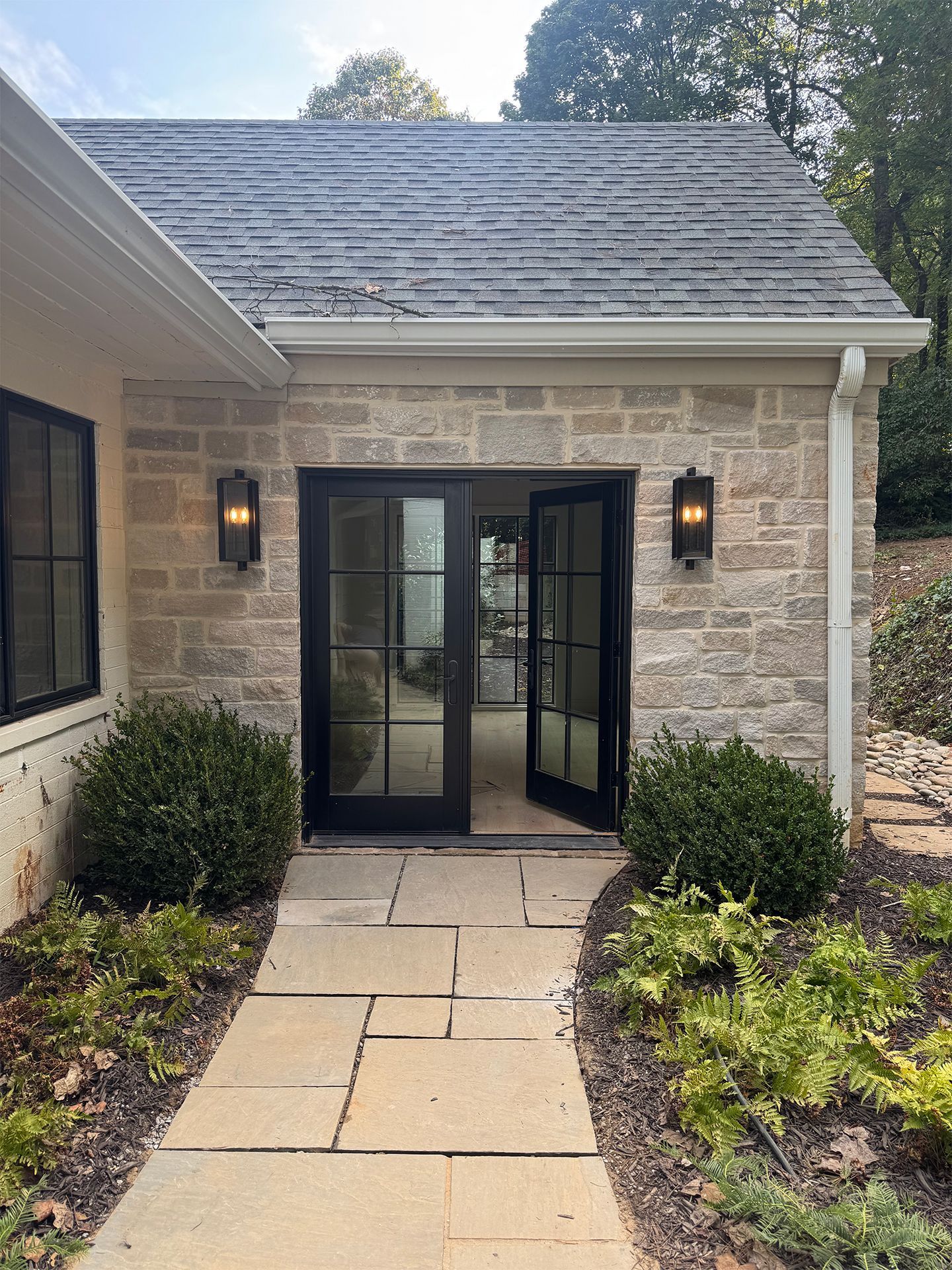 Front entrance of a stone house with black double doors, lit wall lanterns, and a paved walkway lined with shrubs