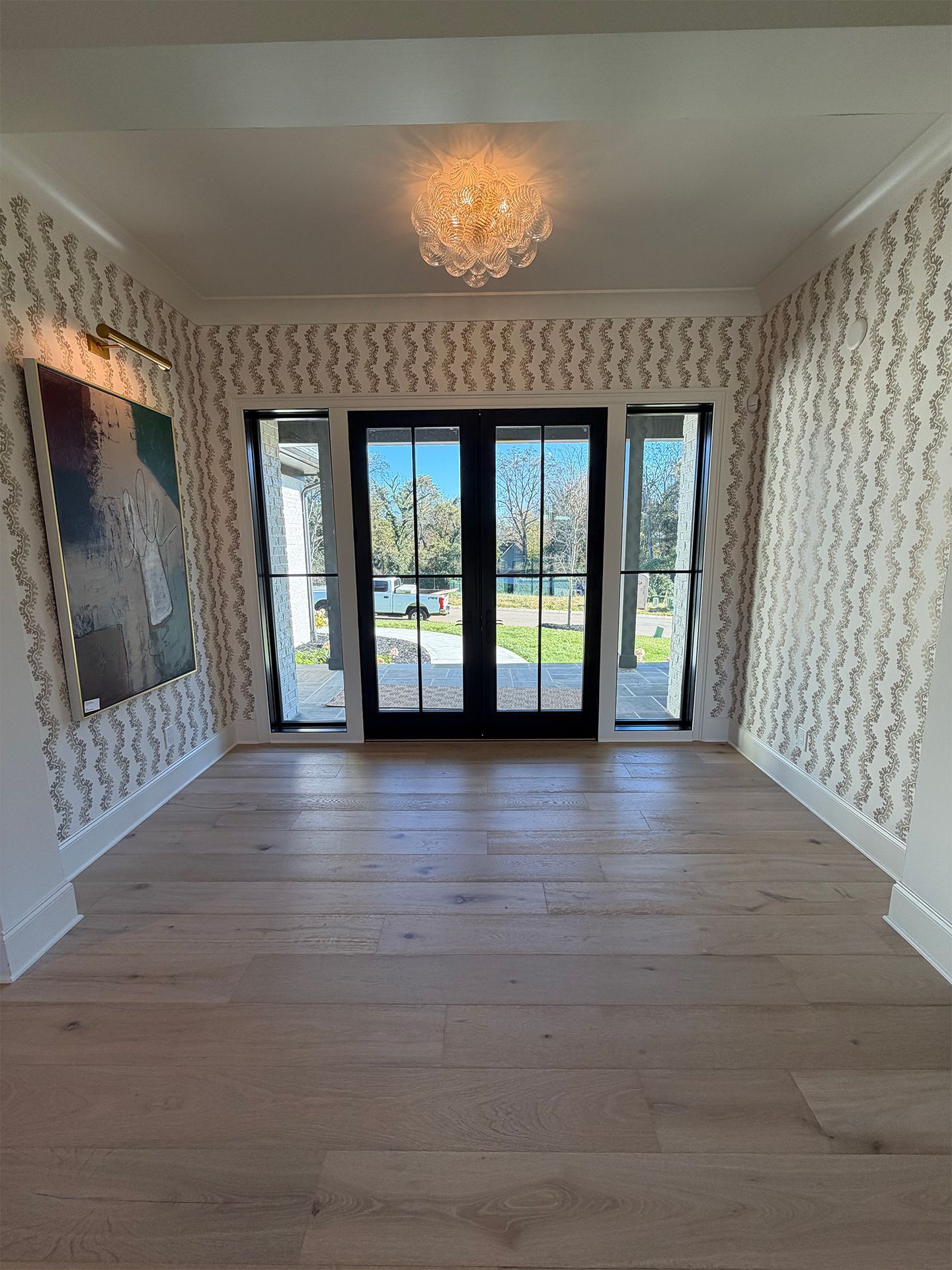 Empty modern room with light wood floors, patterned wallpaper, chandelier, and glass doors to a patio