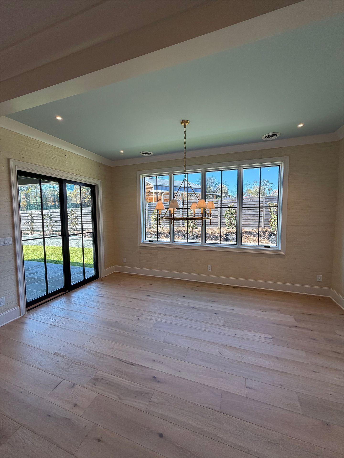 Empty sunlit living room with hardwood floors, large window, and sliding glass doors to a yard.