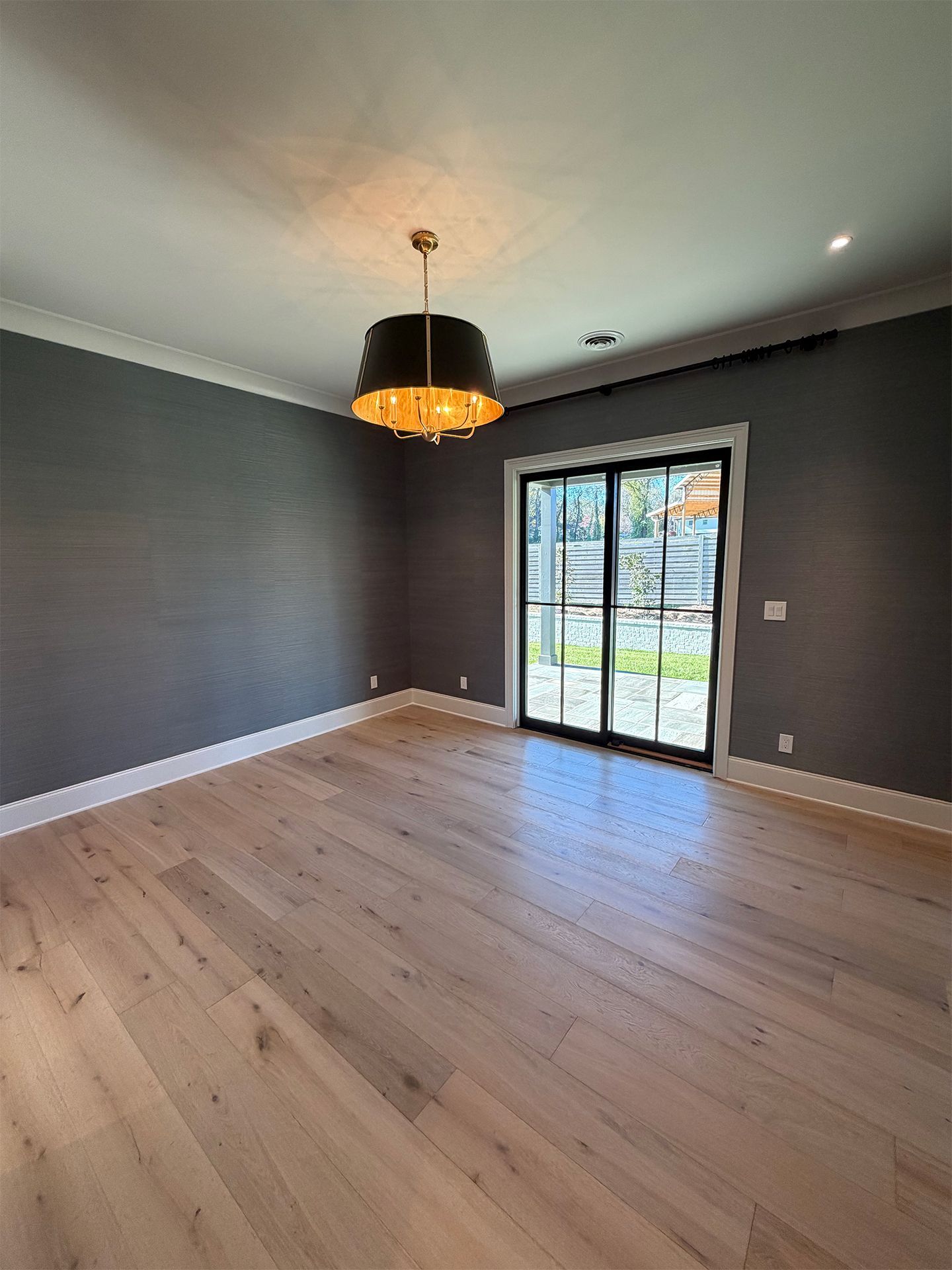 Empty modern room with gray walls, light wood floors, and a black pendant light near a glass door.