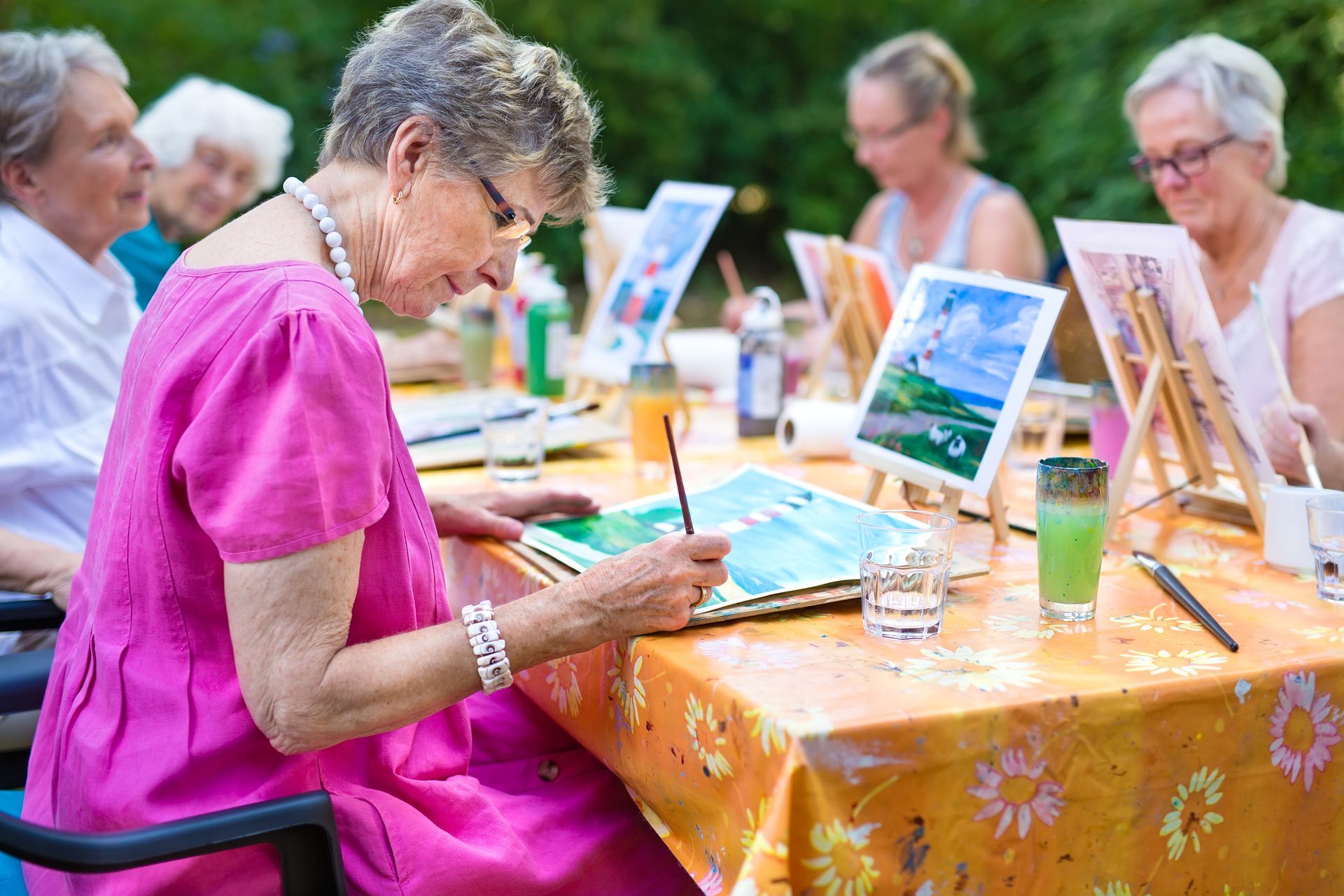 Des personnes sont assises à une table en plein air avec des chevalets, peignant des scènes sur des toiles dans un cadre d'atelier.