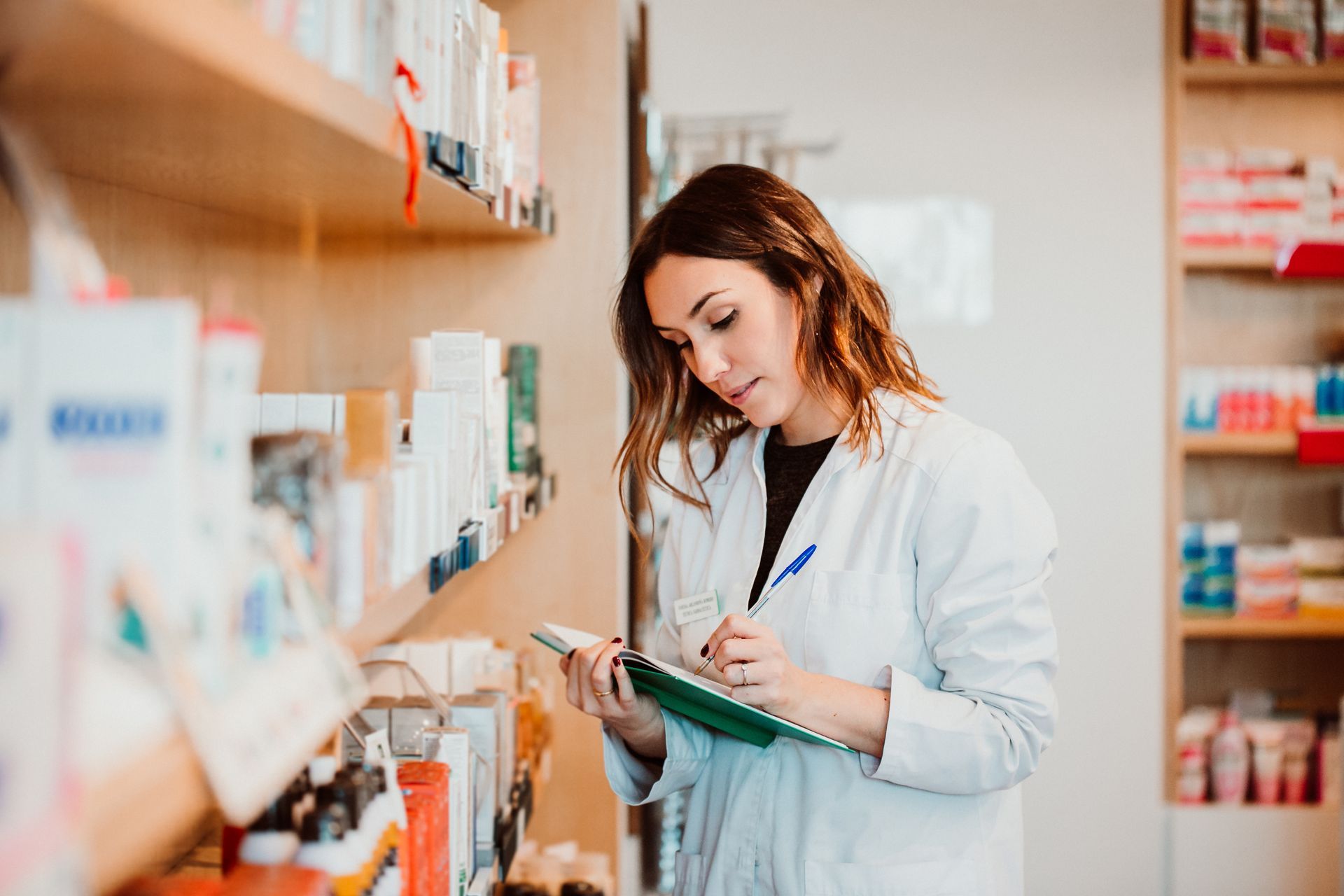 A pharmacist in a white lab coat taking notes on a clipboard while standing in a pharmacy aisle.