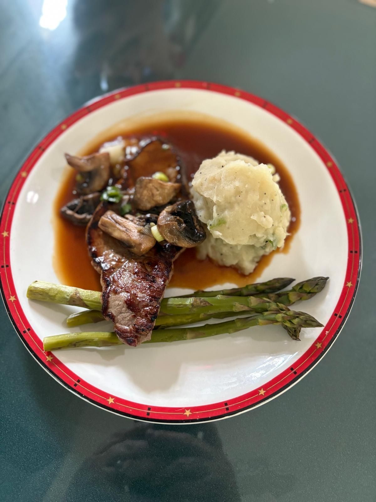 A steak with sautéed mushrooms, brown sauce, mashed potatoes, and asparagus on a white plate with a red patterned rim.