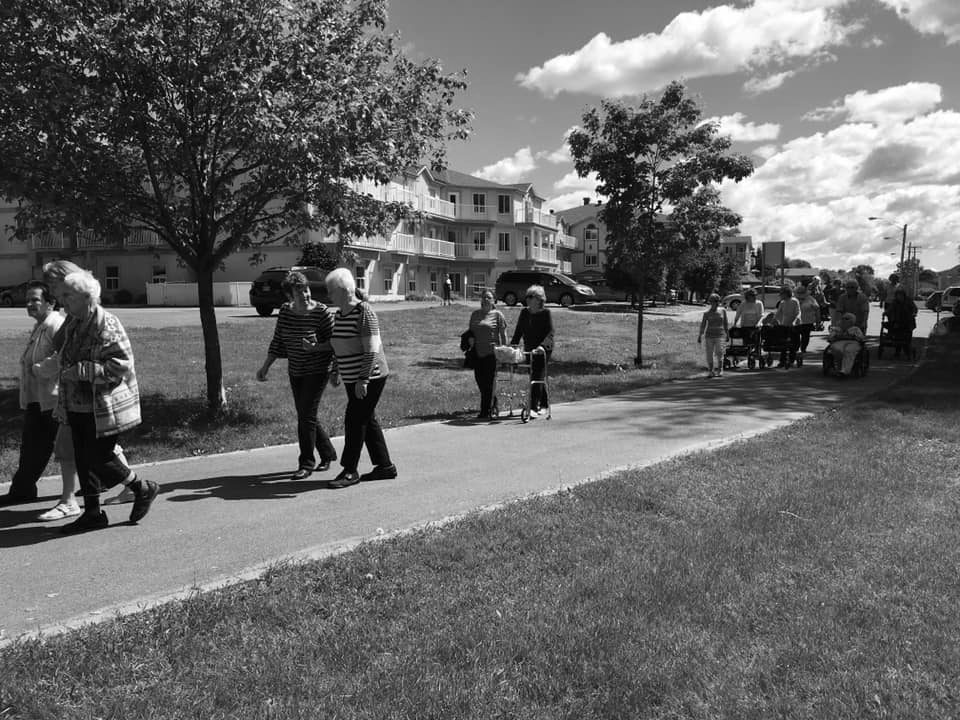 A group of people walking together on a paved path in a park with buildings in the background under a cloudy sky.