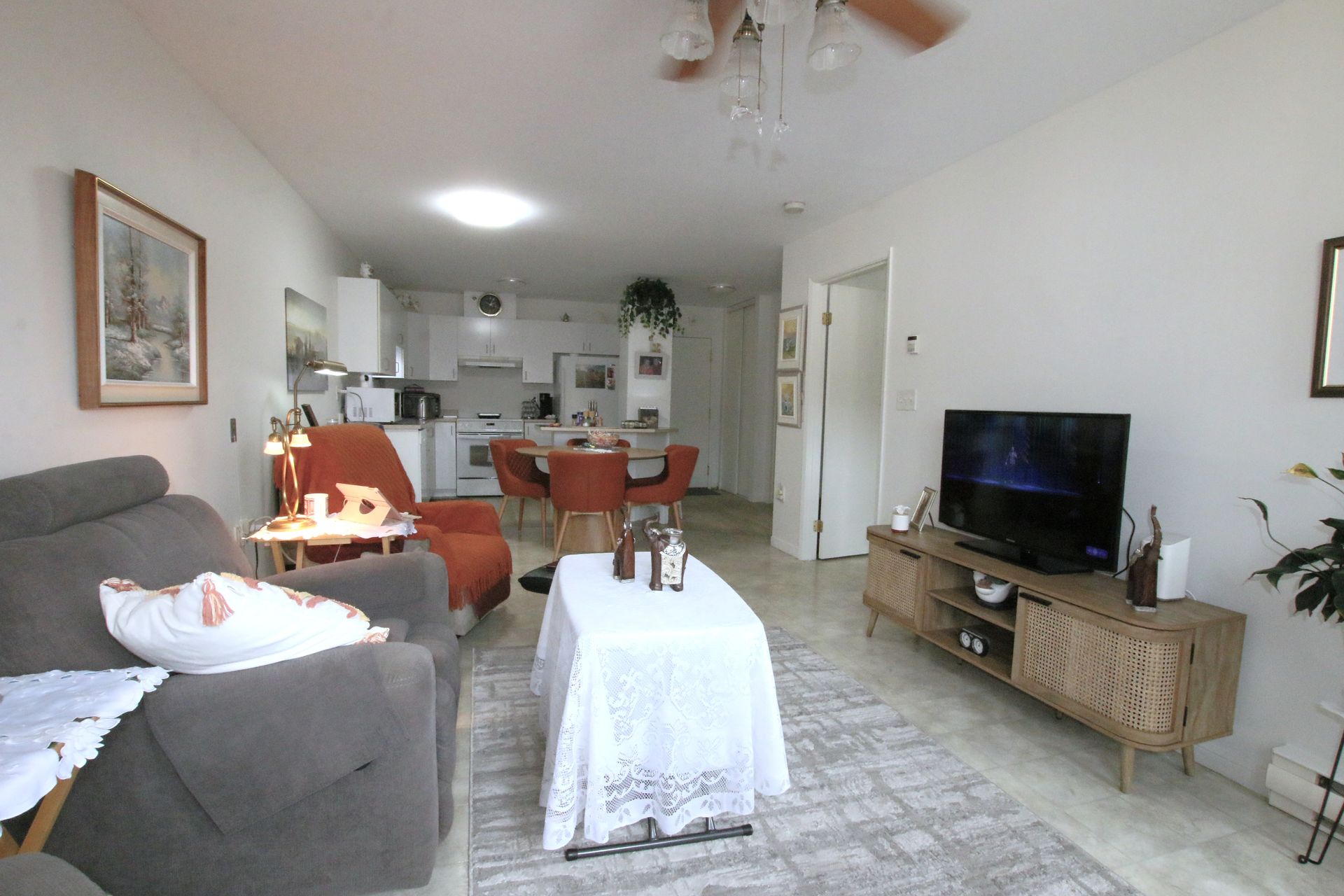 A living room with a gray sofa, a TV on a wooden stand, and an open-concept dining area with a table and chairs in the back.