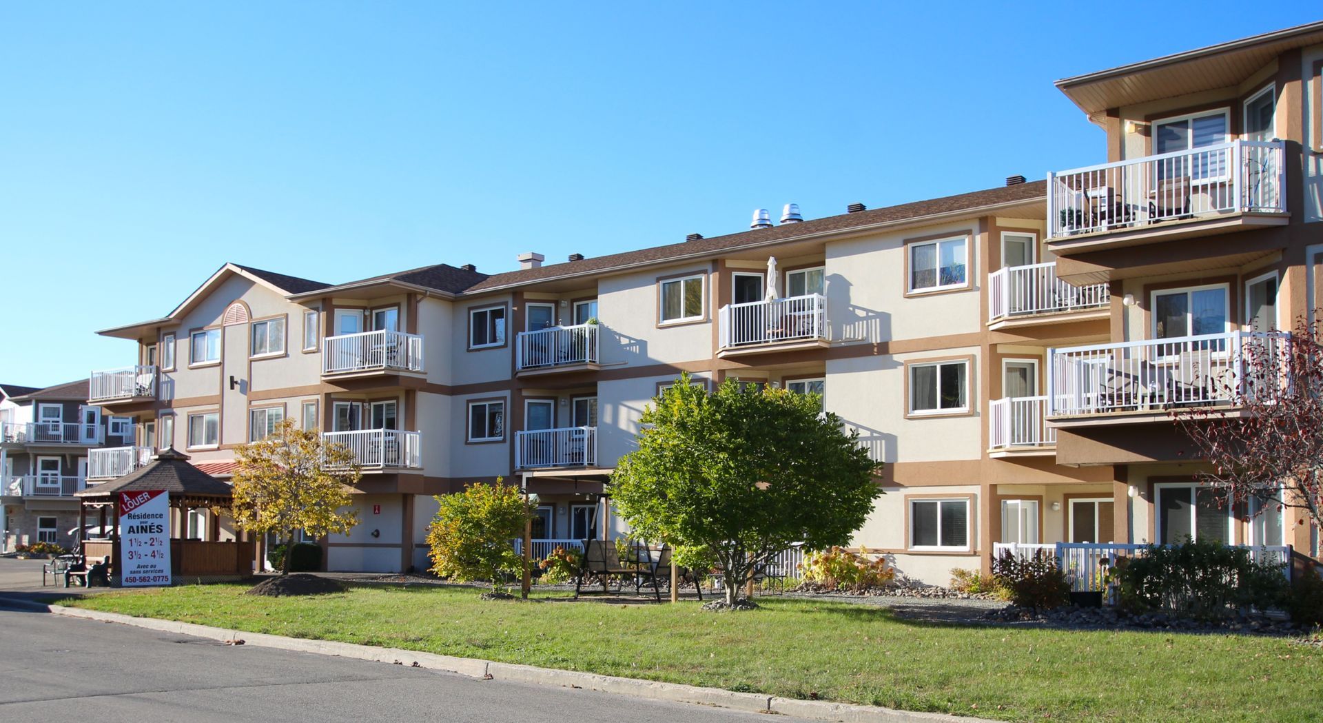A multi-story apartment building with beige siding, white balconies, and small trees on a lawn under a blue sky.