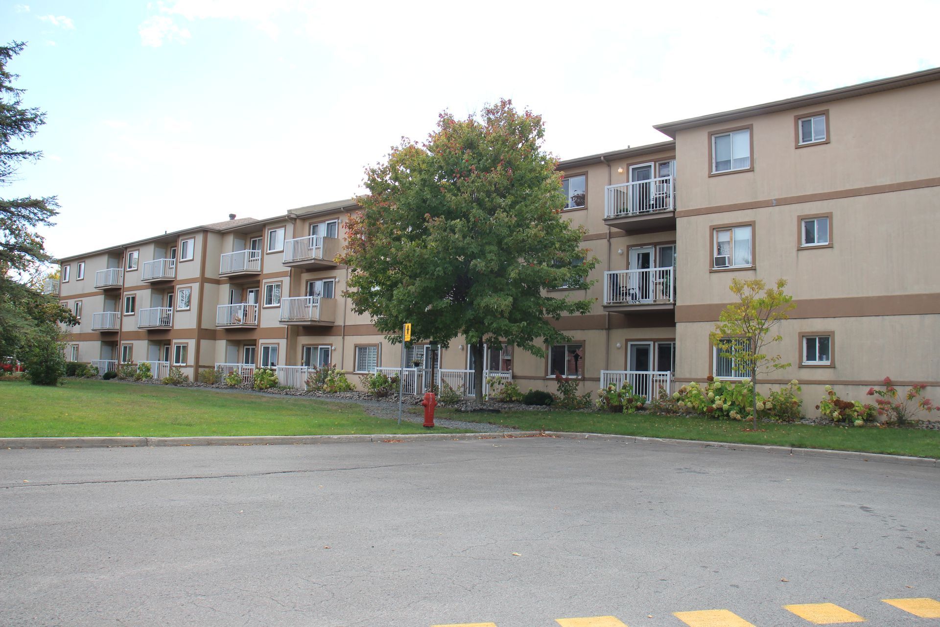 A three-story, beige apartment building with balconies, surrounded by green lawns and a paved parking area.