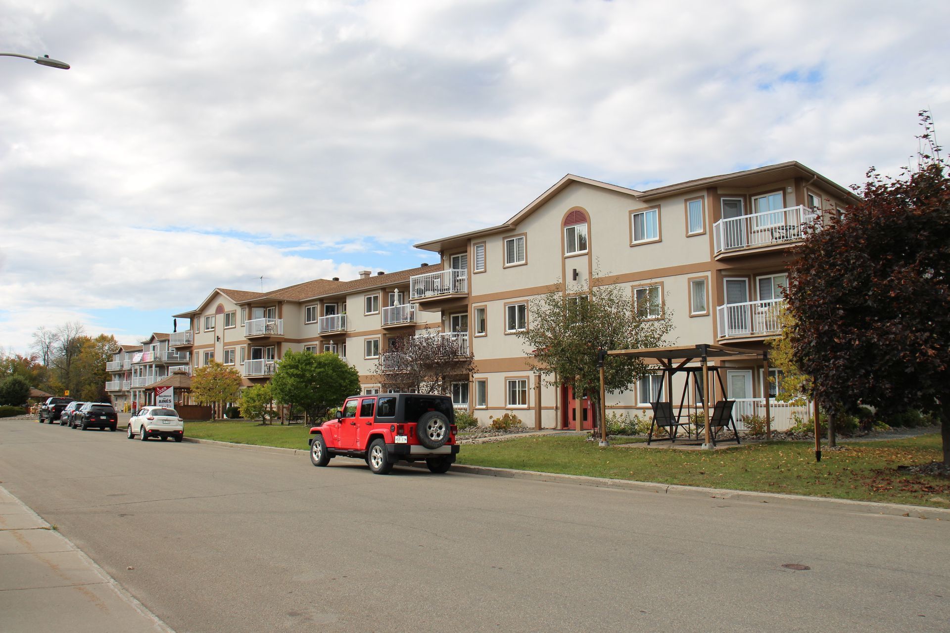 A street-level view of a multi-story apartment building with beige walls and a red Jeep parked in the foreground.