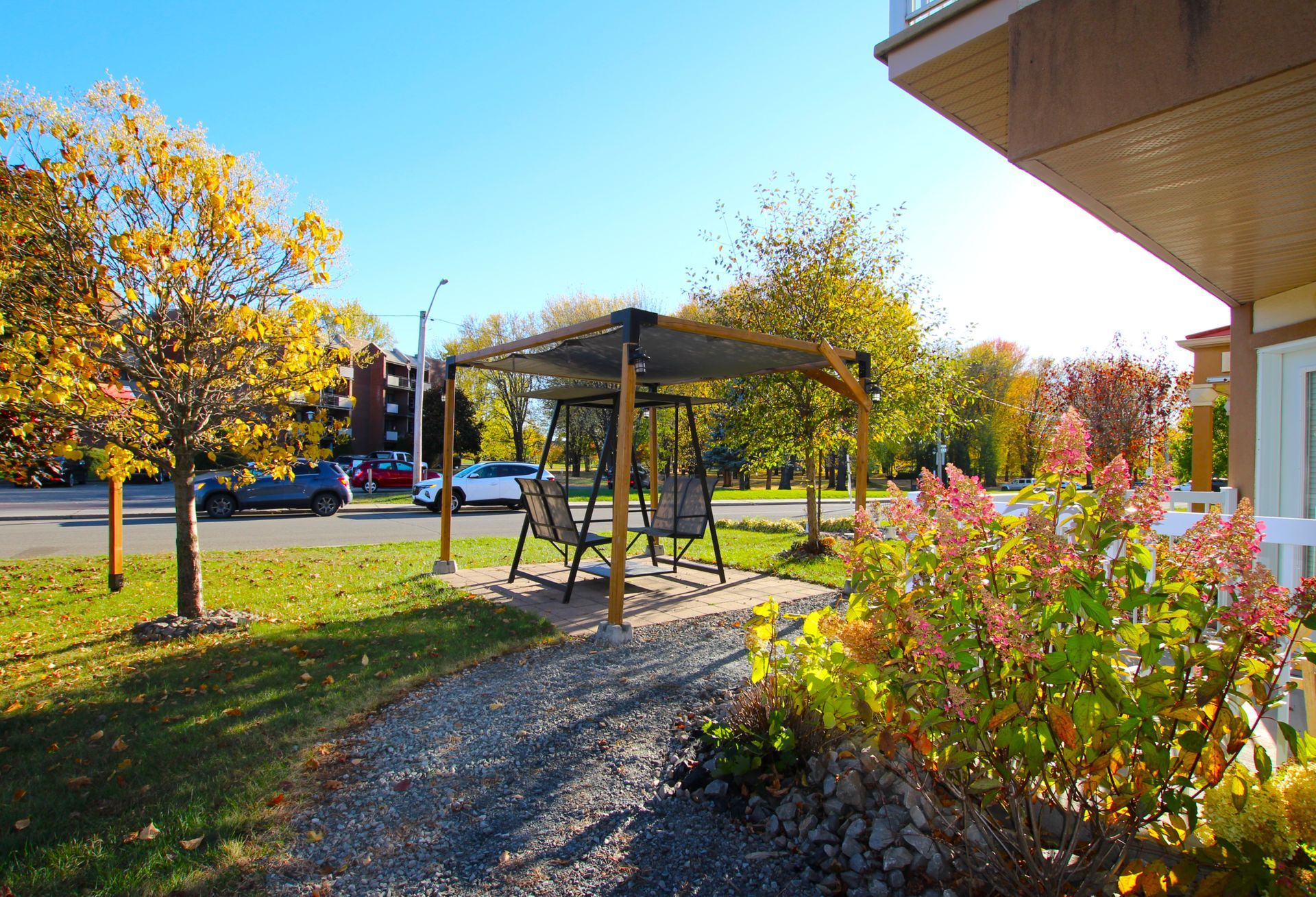 A metal gazebo with a hanging swing on a gravel path near autumn trees and a building exterior under a clear blue sky.