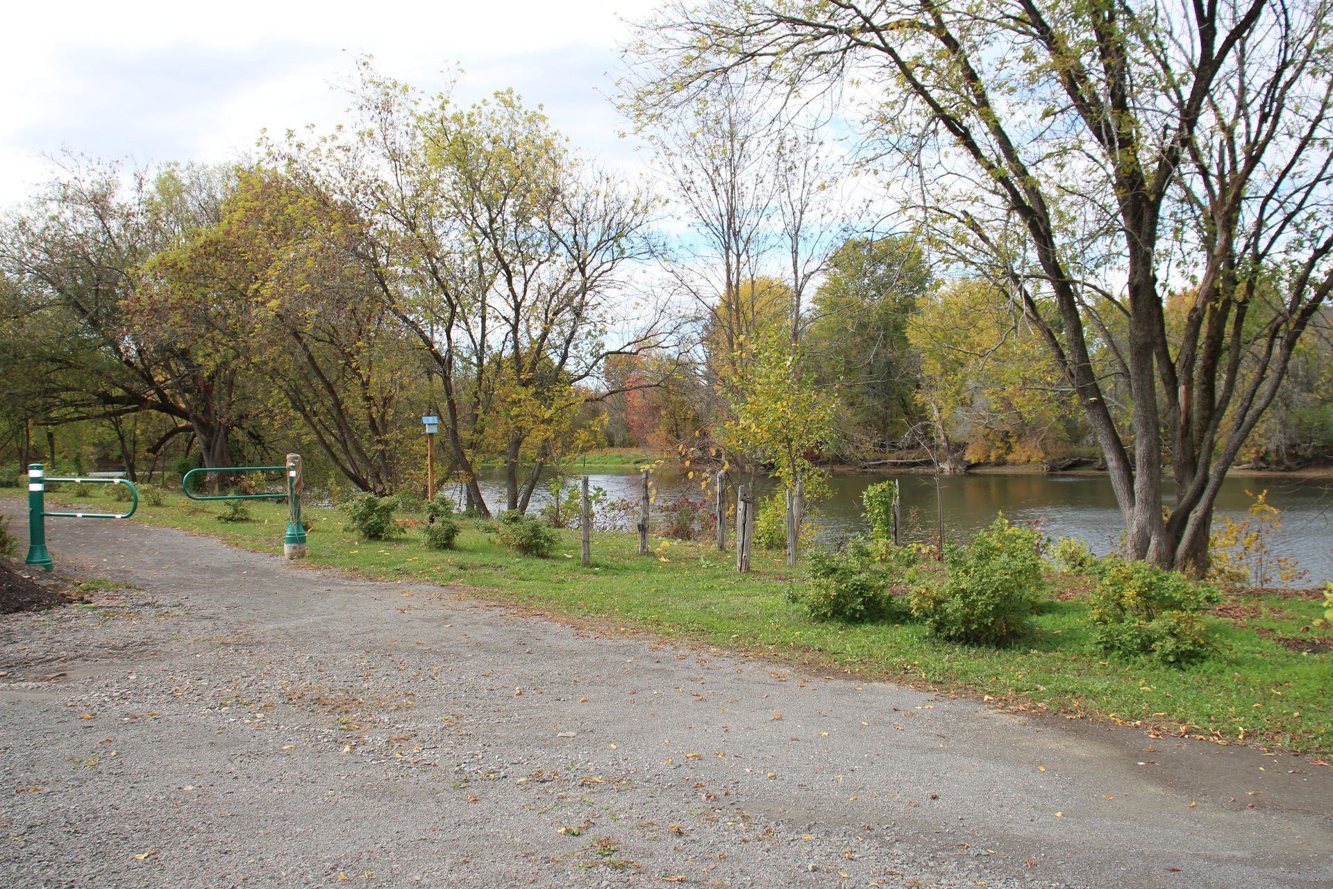 A gravel path borders a calm river, lined with green and autumn-colored trees under a cloudy sky.