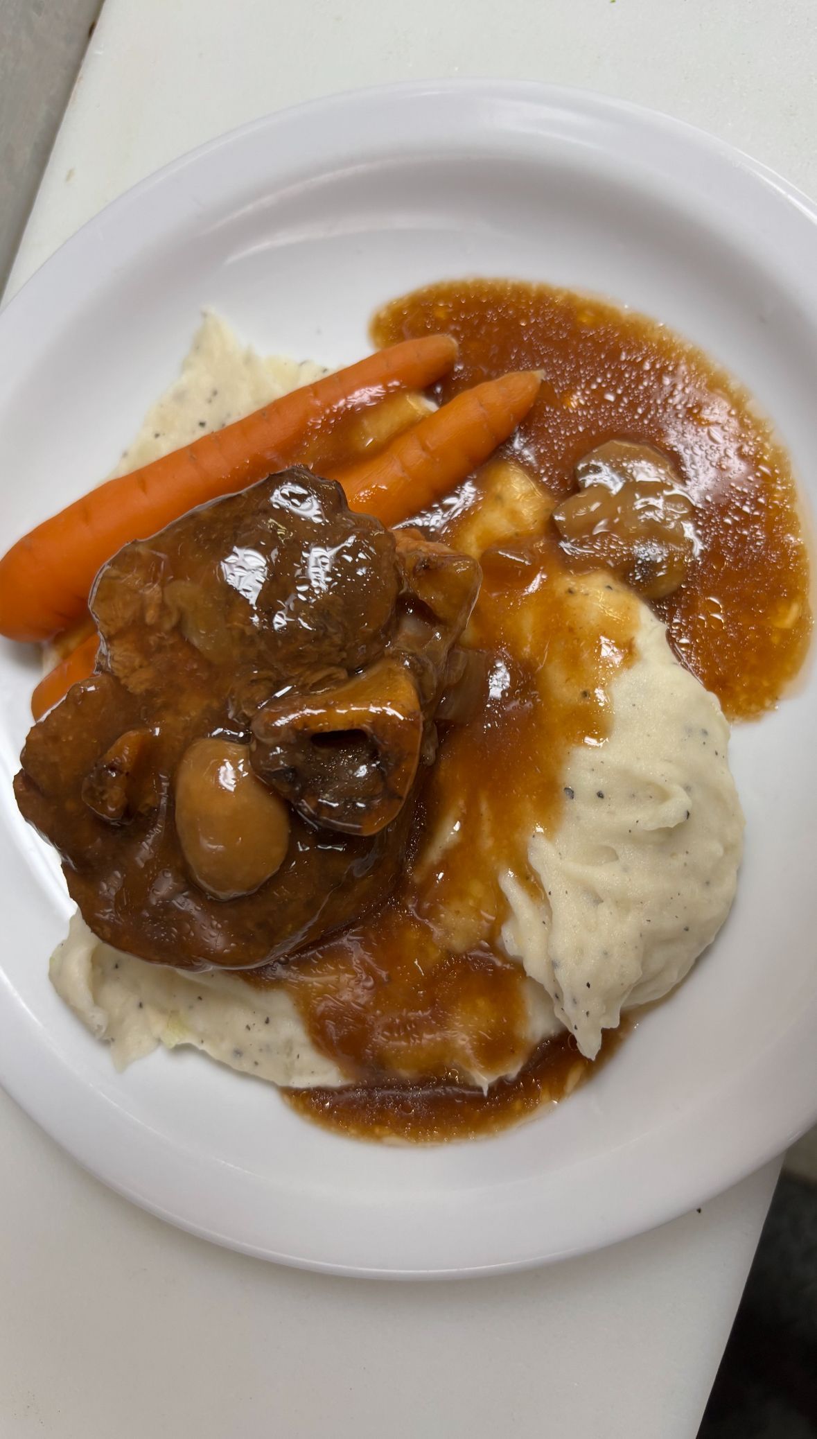 A plated meal featuring braised meat, two whole carrots, and a mound of mashed potatoes covered in brown gravy.